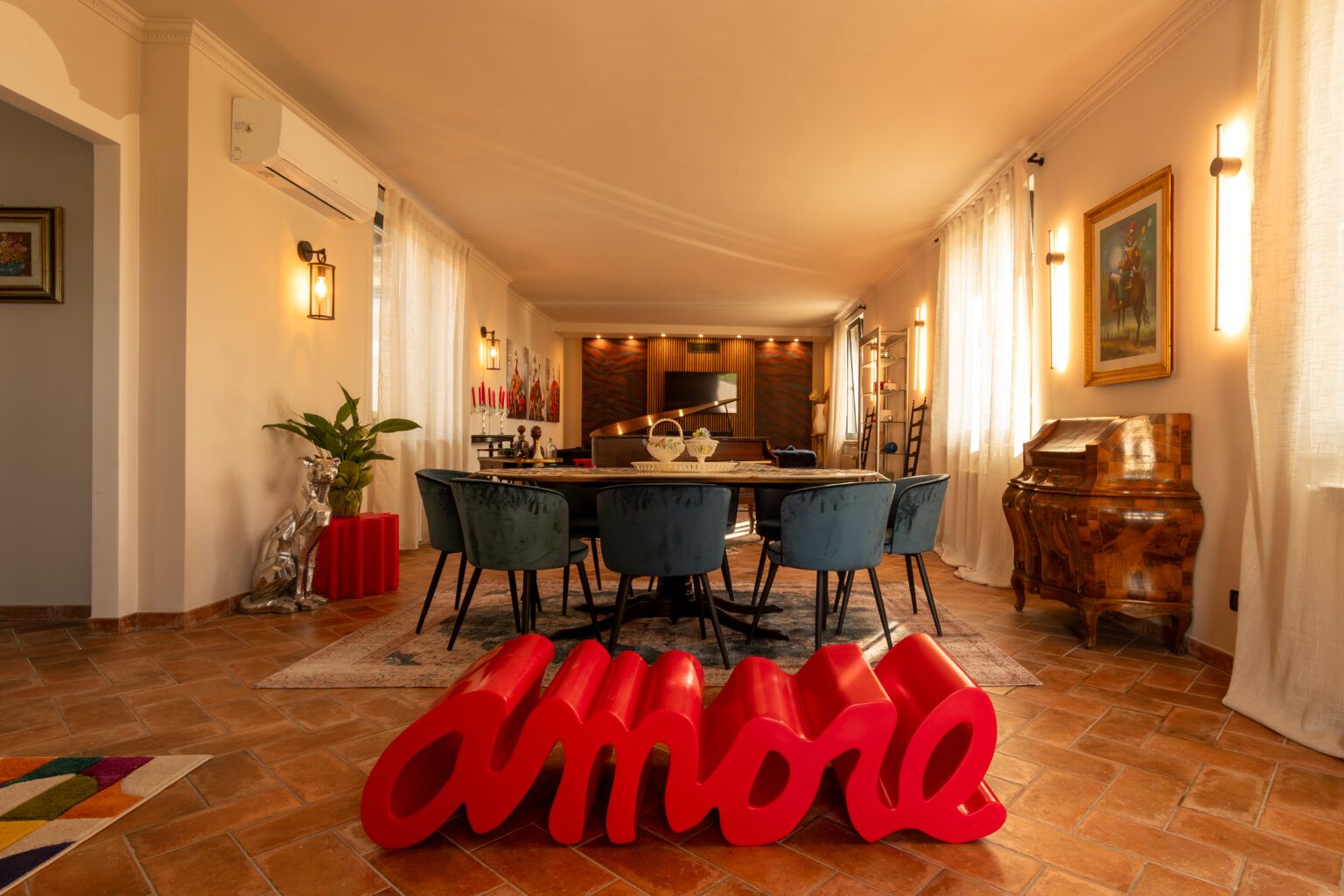 Spacious and stylish dining area featuring a large wooden table surrounded by plush blue chairs, illuminated by natural light from large windows. A vibrant red sculpture spelling "amore" adds a pop of color to the terracotta floor, complemented by decorative elements such as a vintage piano and artwork on the walls. Lush greenery enhances the inviting atmosphere. | Only on SopranoVillas
