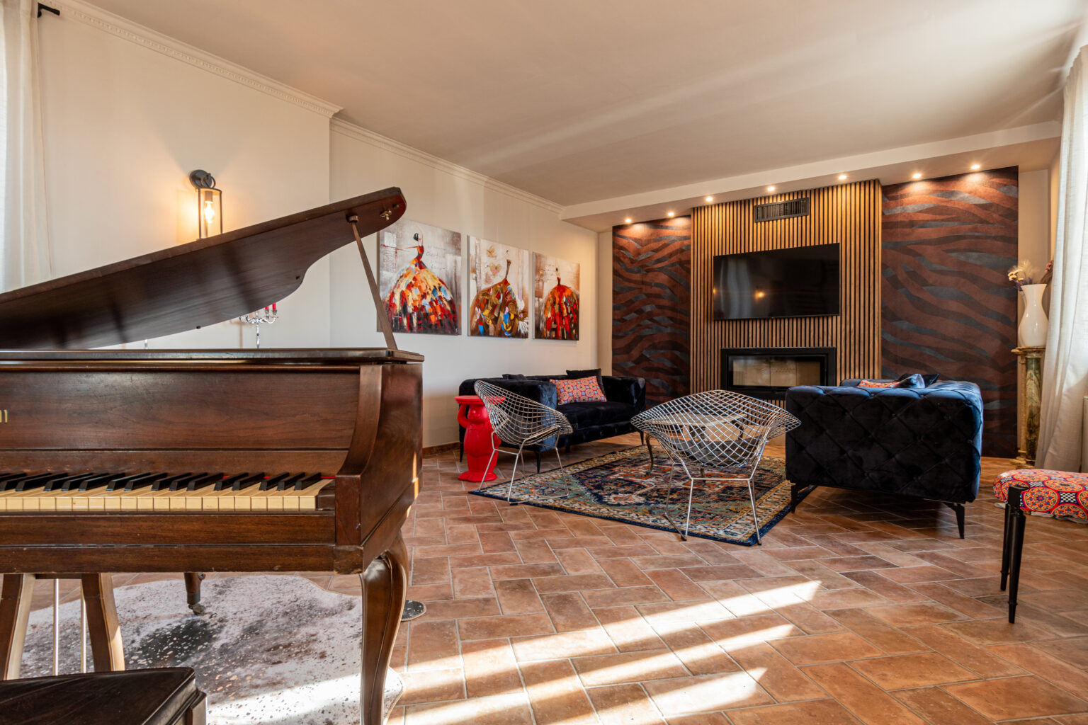 Elegant living room featuring a grand piano, modern art on the walls, plush velvet furniture, and stylish wire chairs. The room has a warm ambiance with natural light, decorative rugs, and a contemporary fireplace. | Only on SopranoVillas