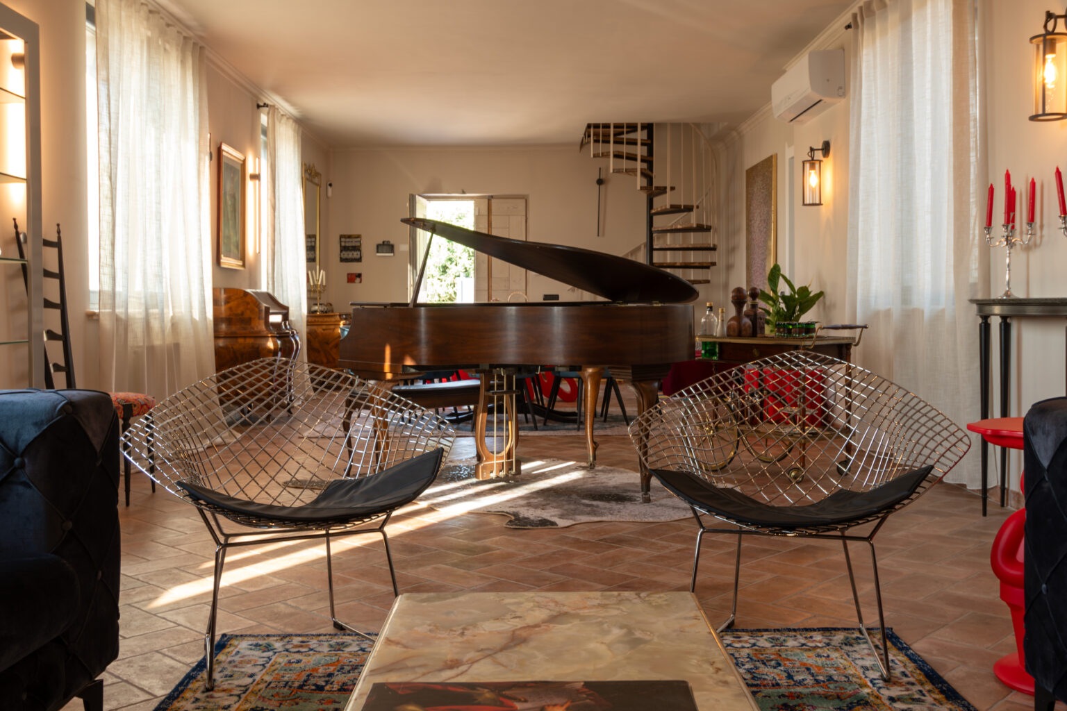Stylish interior of a cozy living space featuring a grand piano, modern wire chairs, and large windows with sheer curtains. The room is adorned with decorative plants, a patterned rug, and artistic elements, creating an inviting atmosphere for relaxation and music. | Only on SopranoVillas
