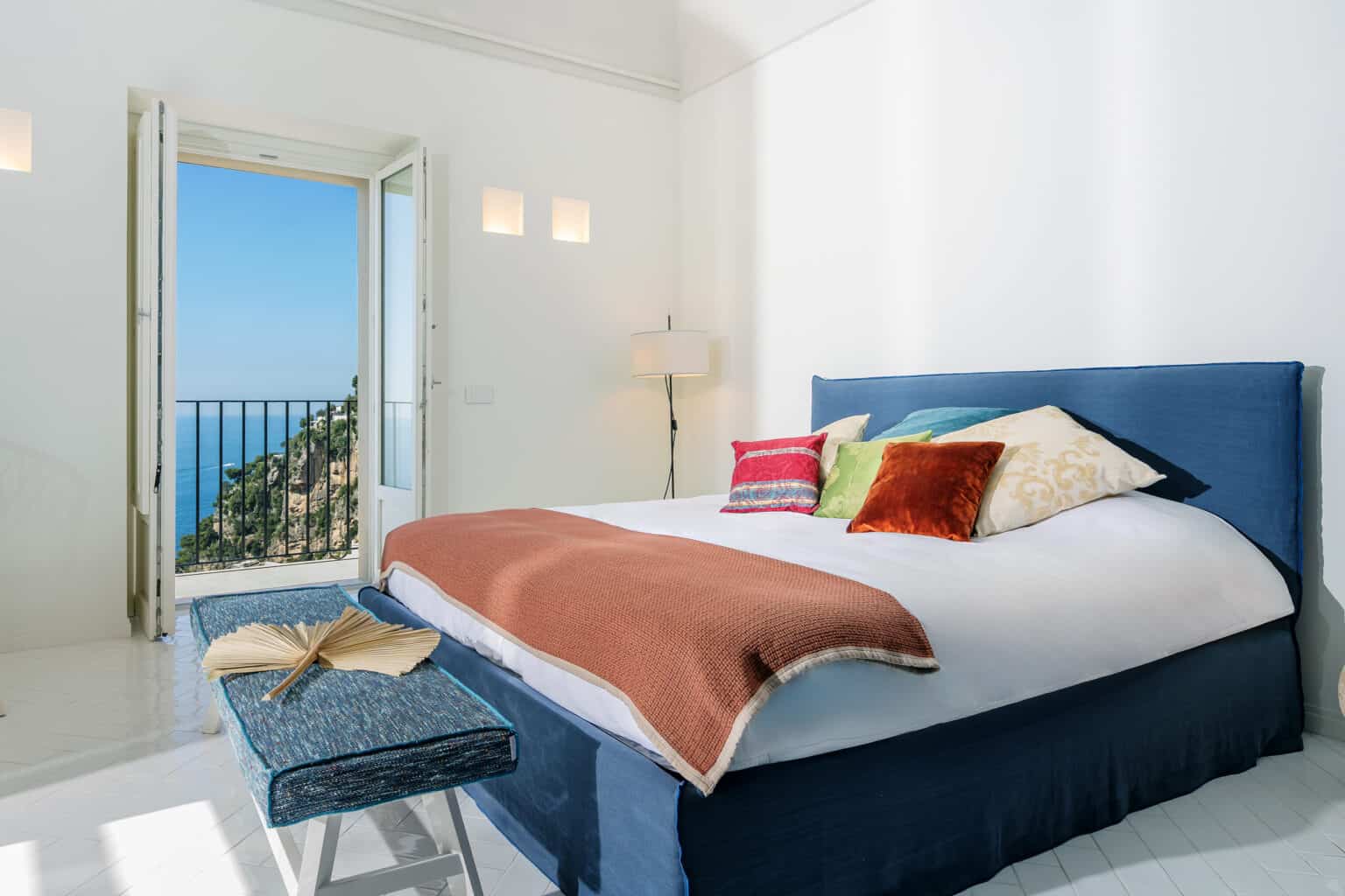 Bright and airy bedroom featuring a large bed adorned with colorful pillows, a cozy blanket, and a stylish bench. The open door reveals a stunning ocean view, enhancing the room's serene atmosphere. Ideal for relaxation and comfort. | Only on SopranoVillas
