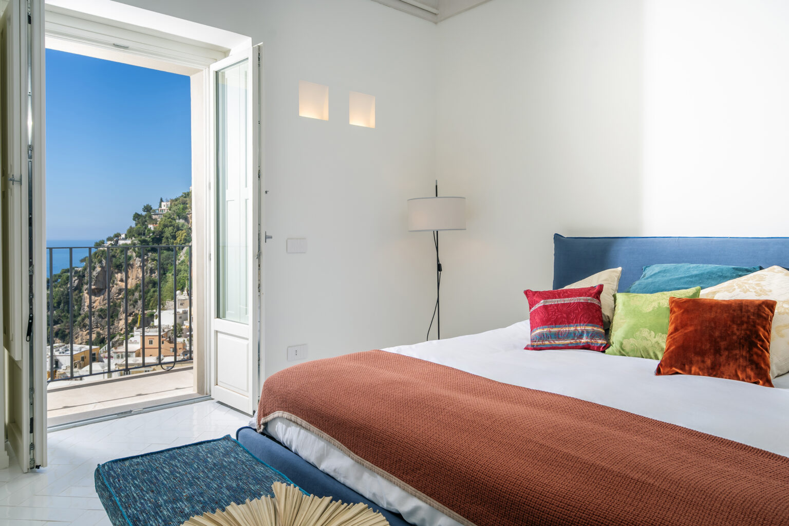 Bright and modern bedroom featuring a large window with a view of the coastline, decorated with colorful pillows on a neatly made bed, a cozy orange throw blanket, and a stylish lamp beside the bed. The room has a serene atmosphere with white walls and elegant furnishings, perfect for relaxation. | Only on SopranoVillas