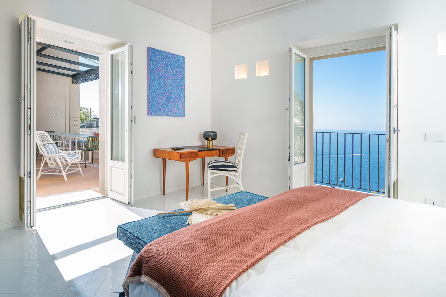 Bright and airy bedroom with a stunning ocean view, featuring a blue accent chair, a wooden desk, and a colorful wall painting. Open doors lead to a balcony with outdoor seating, enhancing the serene atmosphere. Ideal for relaxation and enjoying coastal scenery. | Only on SopranoVillas