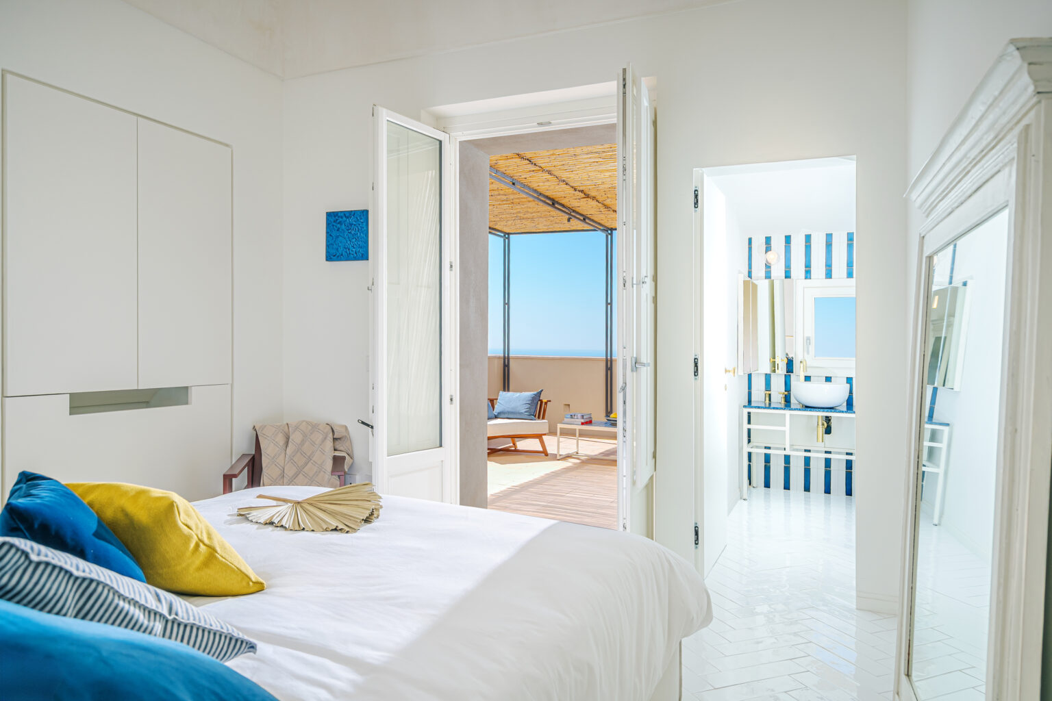 Bright and airy bedroom with a large bed adorned with colorful pillows, featuring an open door leading to a sunlit terrace. The interior includes a modern bathroom with blue and white striped tiles and a stylish sink. Natural light floods the space, highlighting the minimalist decor and serene atmosphere. | Only on SopranoVillas