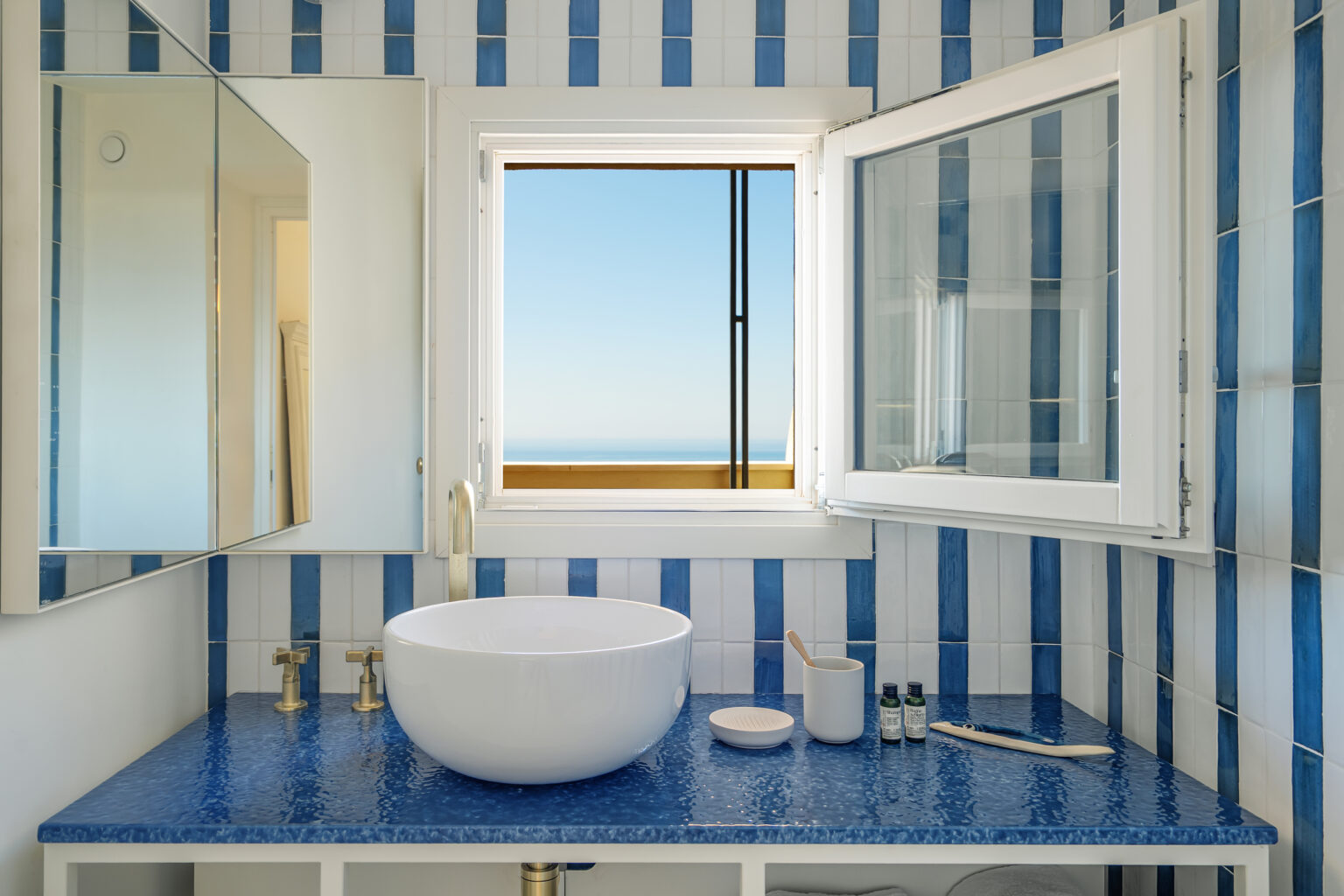 Modern bathroom with blue and white striped tiles, featuring a round white sink, gold faucets, and a window offering a scenic ocean view. The countertop is adorned with toiletries and a mirror, creating a stylish and serene atmosphere. | Only on SopranoVillas