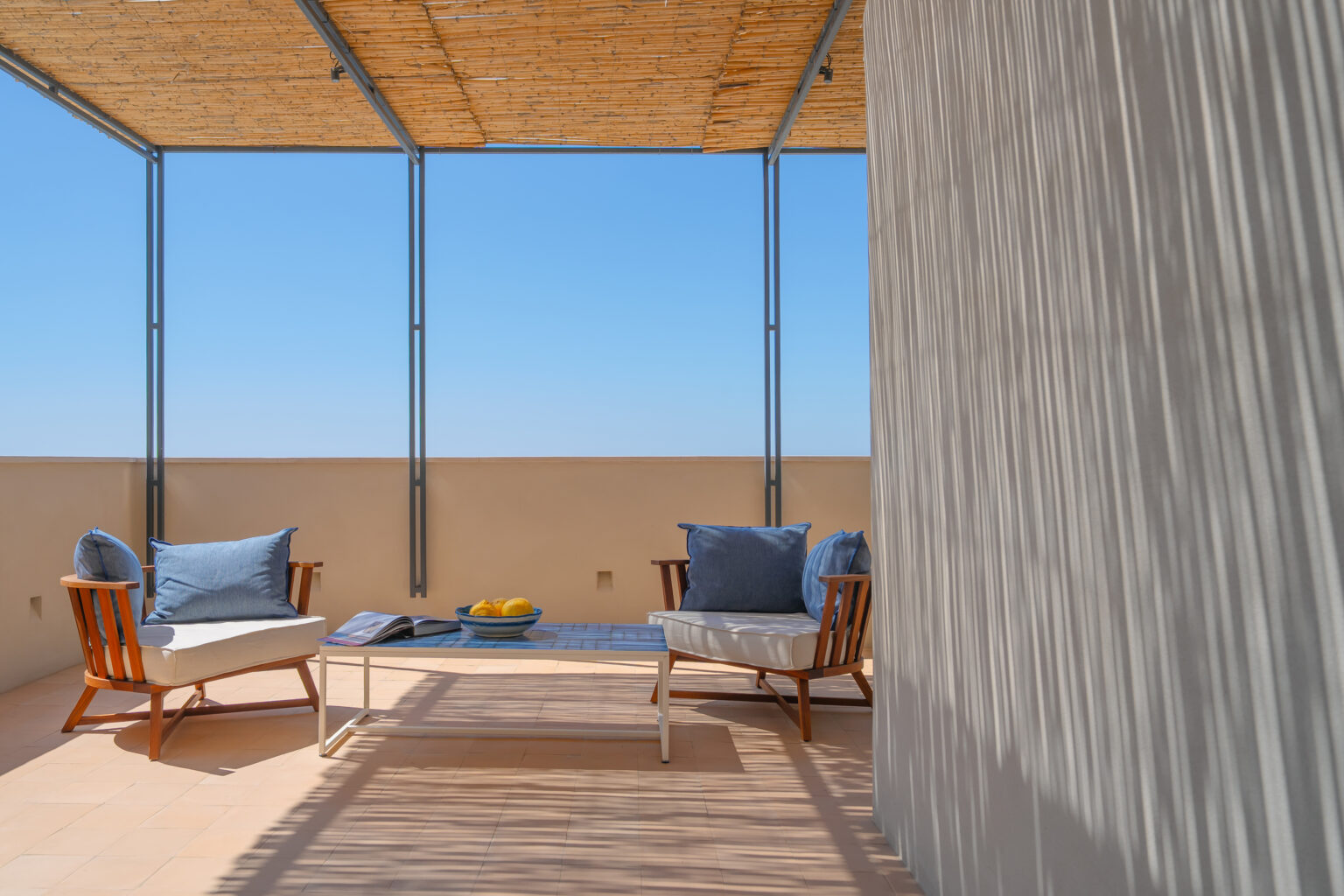 Modern outdoor terrace with two wooden chairs and blue cushions, a glass coffee table with a bowl of lemons, and a clear blue sky above. Natural light creates a relaxing atmosphere, ideal for enjoying the outdoors. | Only on SopranoVillas