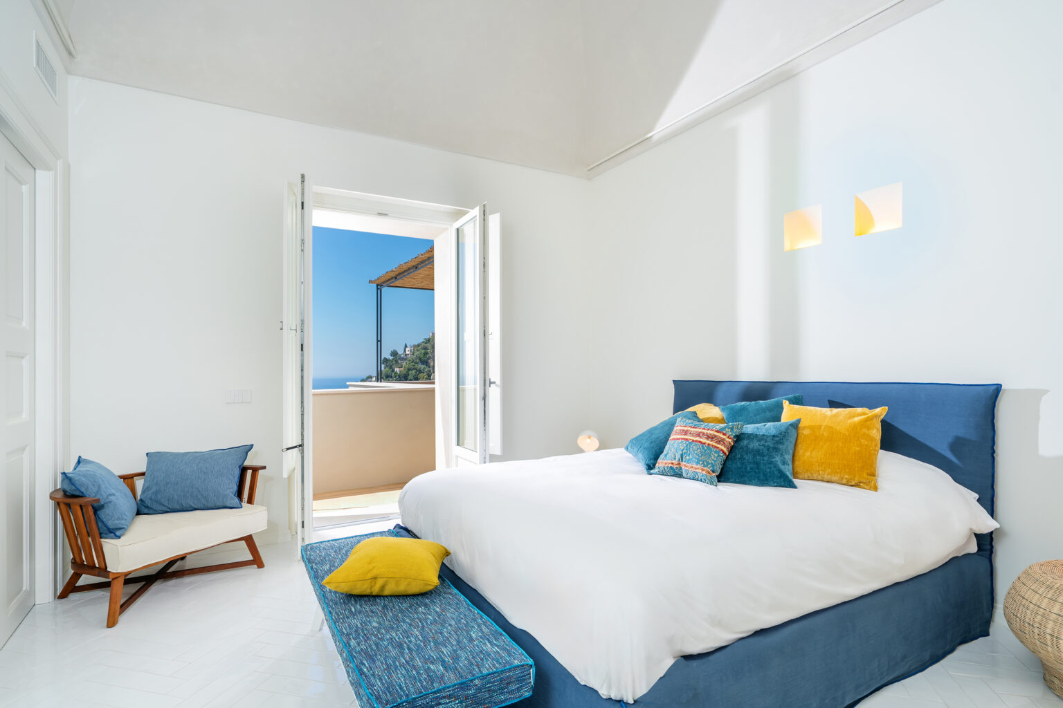 Bright and airy bedroom featuring a large bed adorned with colorful pillows, a cozy seating area with a blue accent chair, and an open balcony door showcasing a scenic view. Natural light floods the space, highlighting the minimalist decor and white walls. | Only on SopranoVillas