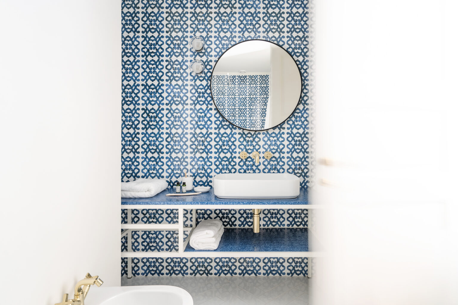 Modern bathroom featuring a blue and white patterned wall, a round mirror, a sleek white sink, and neatly arranged towels on a stylish shelf. The design emphasizes a clean and contemporary aesthetic. | Only on SopranoVillas