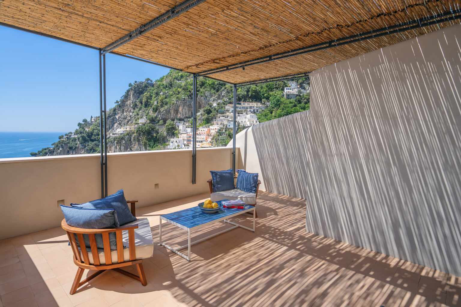 Stylish outdoor terrace with a wooden seating area, blue cushions, and a coffee table, offering stunning views of a coastal landscape and lush hills. Ideal for relaxation and enjoying the Mediterranean climate. | Only on SopranoVillas