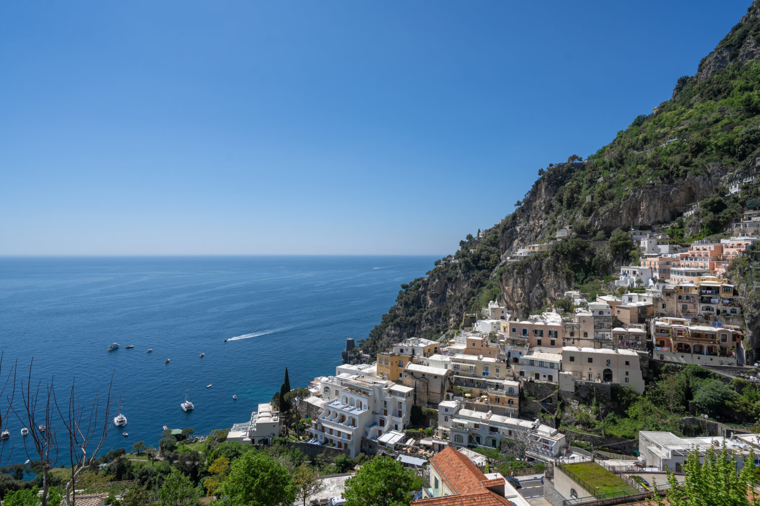 Panoramic view of Positano, Italy, showcasing colorful cliffside homes, lush greenery, and the serene blue waters of the Mediterranean Sea under a clear sky. | Only on SopranoVillas