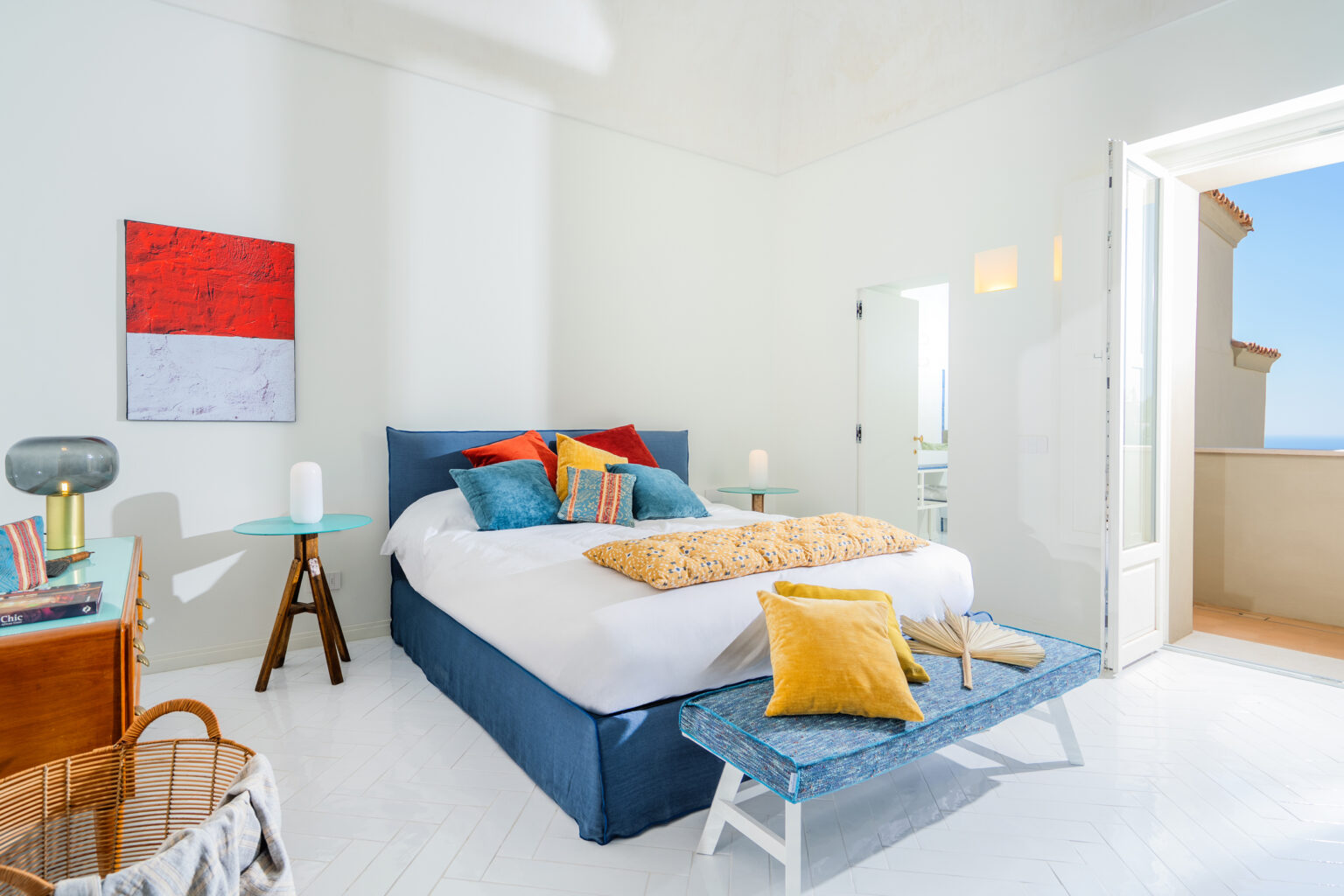 Bright and airy modern bedroom featuring a large bed adorned with colorful pillows, a textured yellow throw, and a stylish blue bed frame. The room includes a wooden accent table, a decorative lamp, and a basket, with an open door leading to a balcony that offers a scenic view. The walls are painted white, enhancing the room's spacious feel. | Only on SopranoVillas