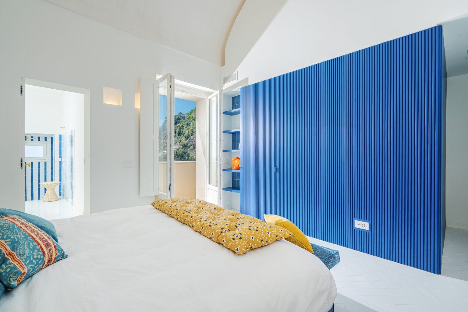 Bright and airy bedroom featuring a large bed with decorative pillows, a vibrant blue accent wall, and open windows that showcase a scenic view. The room includes a modern design with a minimalist aesthetic, natural light flooding in, and a glimpse of a stylish bathroom in the background. | Only on SopranoVillas