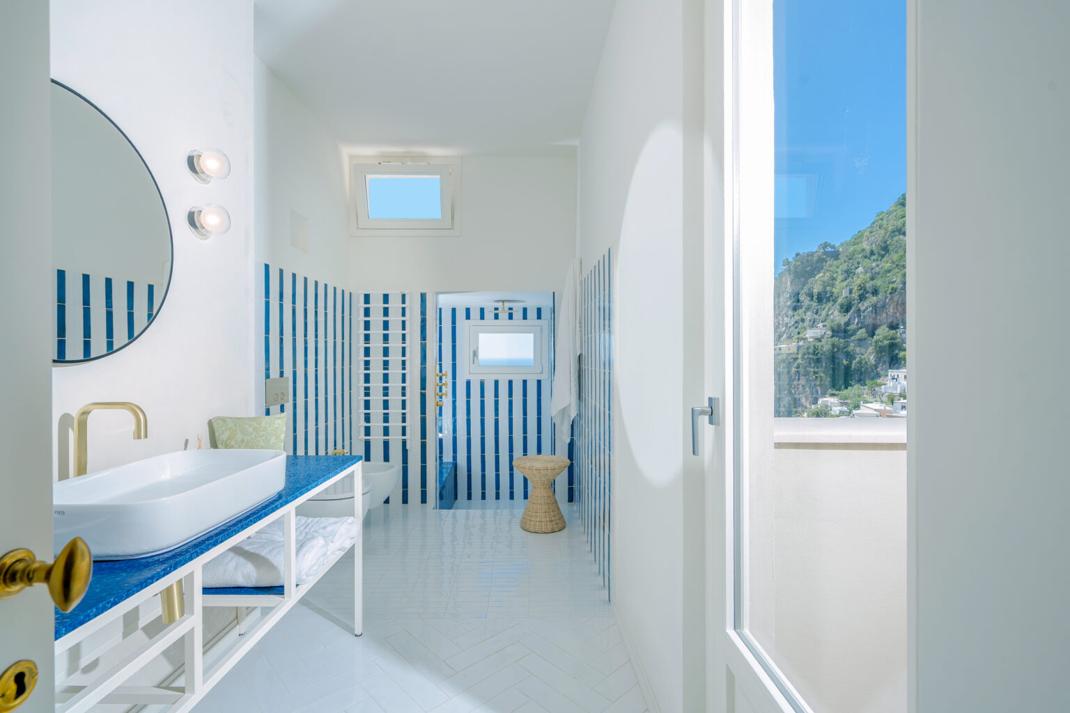 Modern bathroom interior featuring white and blue striped tiles, a circular mirror, a stylish sink on a blue countertop, and natural light streaming through a window. The design emphasizes a clean, airy aesthetic with a view of green hills outside. | Only on SopranoVillas