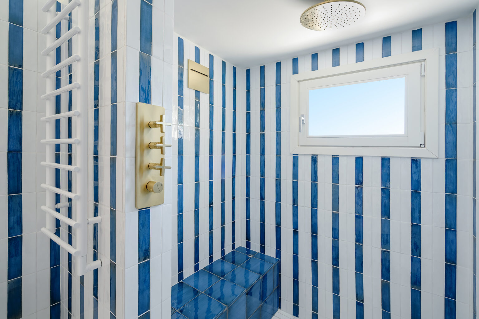Modern bathroom shower featuring blue and white striped tiles, a sleek gold faucet control panel, and a window for natural light. The space includes a built-in bench and a stylish overhead showerhead, creating a contemporary and inviting atmosphere. | Only on SopranoVillas