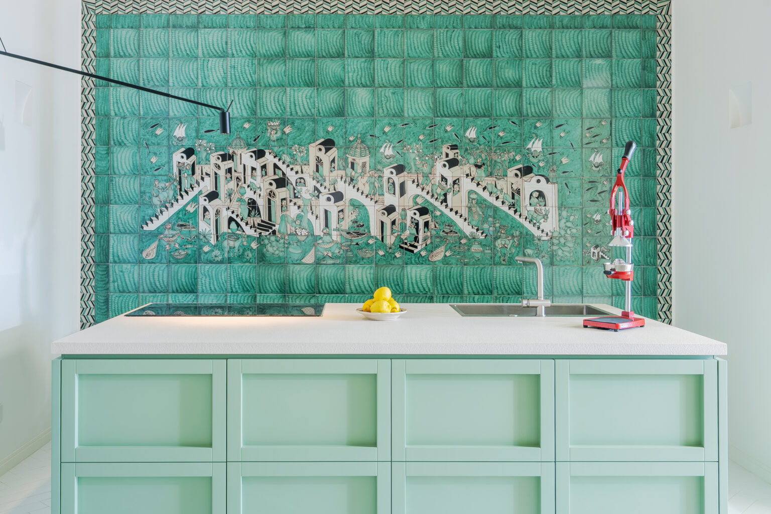 Modern kitchen featuring a mint green island and artistic tile mural depicting a vibrant cityscape. The space includes a sleek sink and a red citrus juicer, complemented by fresh lemons in a bowl. | Only on SopranoVillas