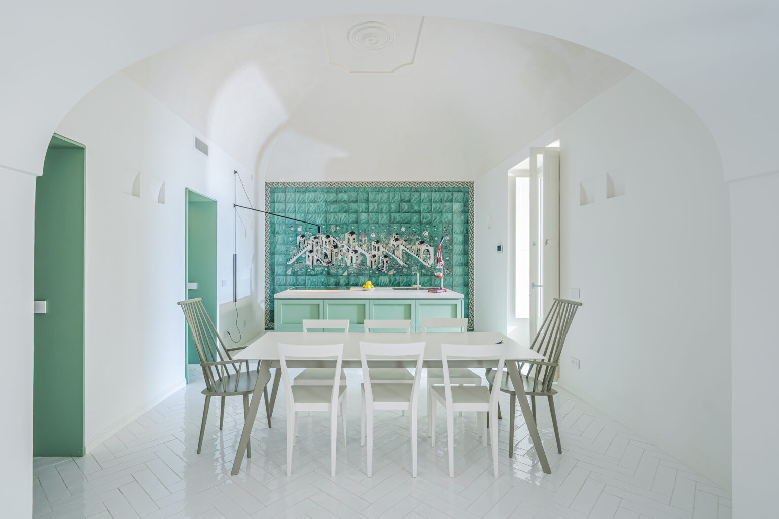 Bright and modern kitchen dining area featuring a green accent wall, white furniture, and sleek design elements. The space includes a minimalist dining table with contemporary chairs, large windows allowing natural light, and a unique decorative backsplash. Ideal for showcasing interior design and home decor trends. | Only on SopranoVillas