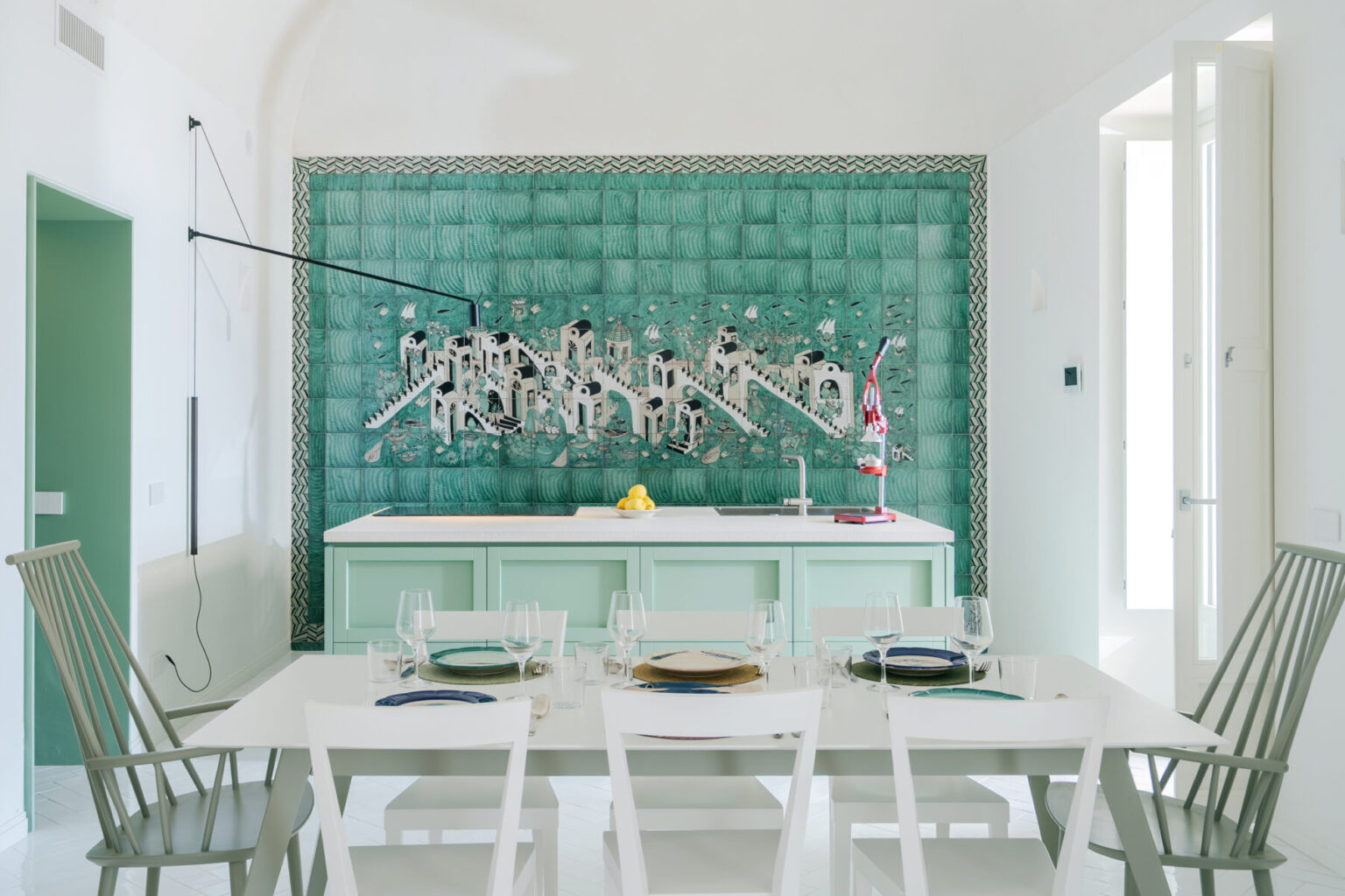 Modern kitchen and dining area featuring a vibrant green tiled wall mural, minimalist white dining table set with tableware, and a sleek kitchen island with a white countertop. The space is well-lit and showcases contemporary design elements. | Only on SopranoVillas
