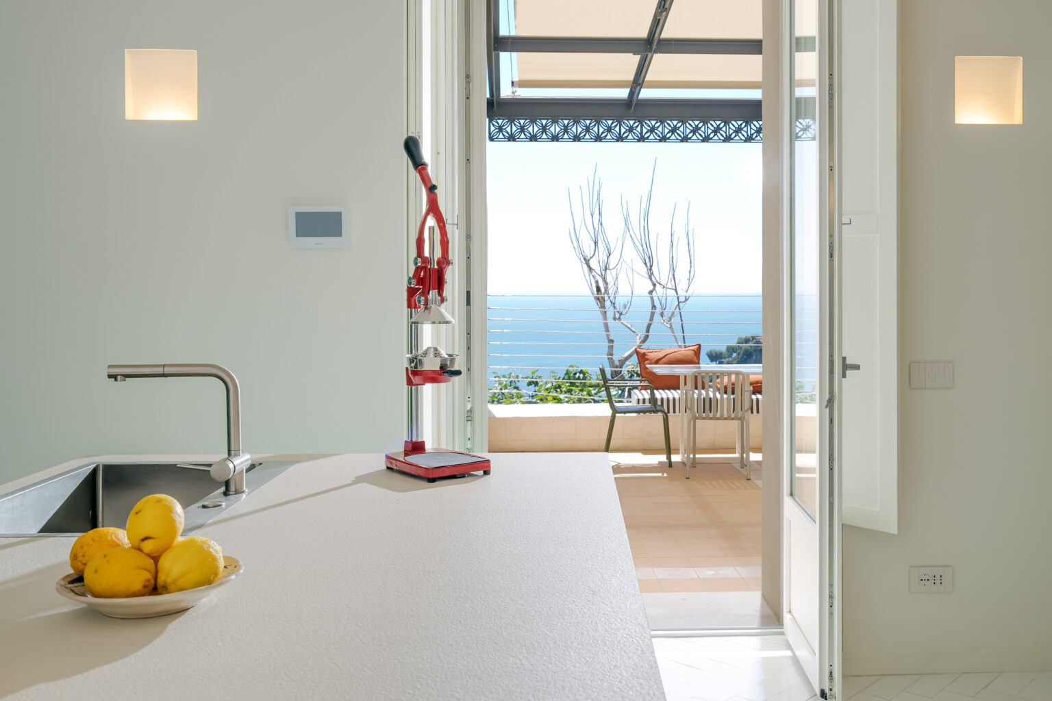 Modern kitchen with a view featuring a sleek countertop, stainless steel sink, and a red citrus juicer. A bowl of lemons sits on the counter, while large doors open to a terrace overlooking the ocean and a minimalist outdoor seating area. Bright, natural light enhances the airy atmosphere. | Only on SopranoVillas