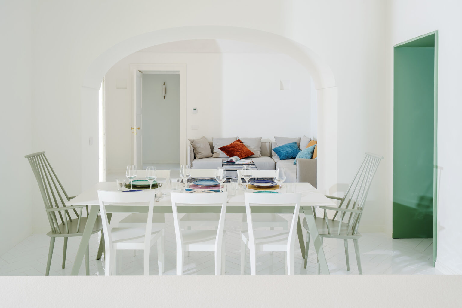 Modern dining area featuring a white table set with colorful plates and glassware, surrounded by stylish green and white chairs. In the background, a cozy living space with a sofa adorned with vibrant cushions is visible through an arched entryway. The room is bright and minimalist with a light color palette. | Only on SopranoVillas