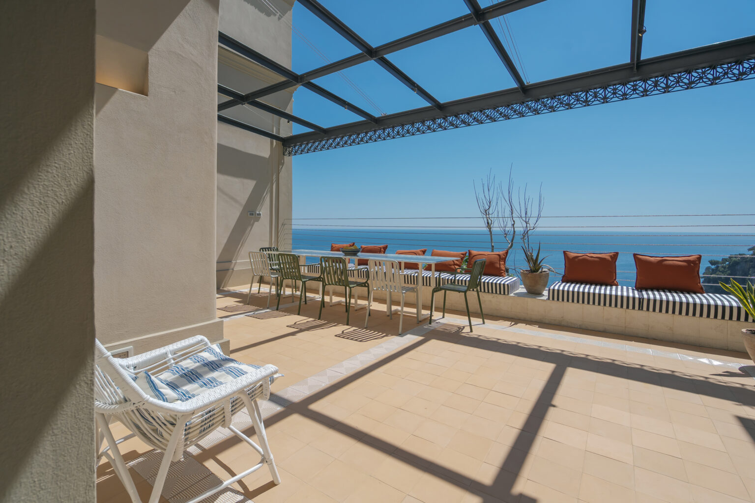 Spacious terrace with ocean view, featuring a long dining table, colorful cushions, and stylish seating under a glass canopy, perfect for outdoor gatherings and relaxation. | Only on SopranoVillas