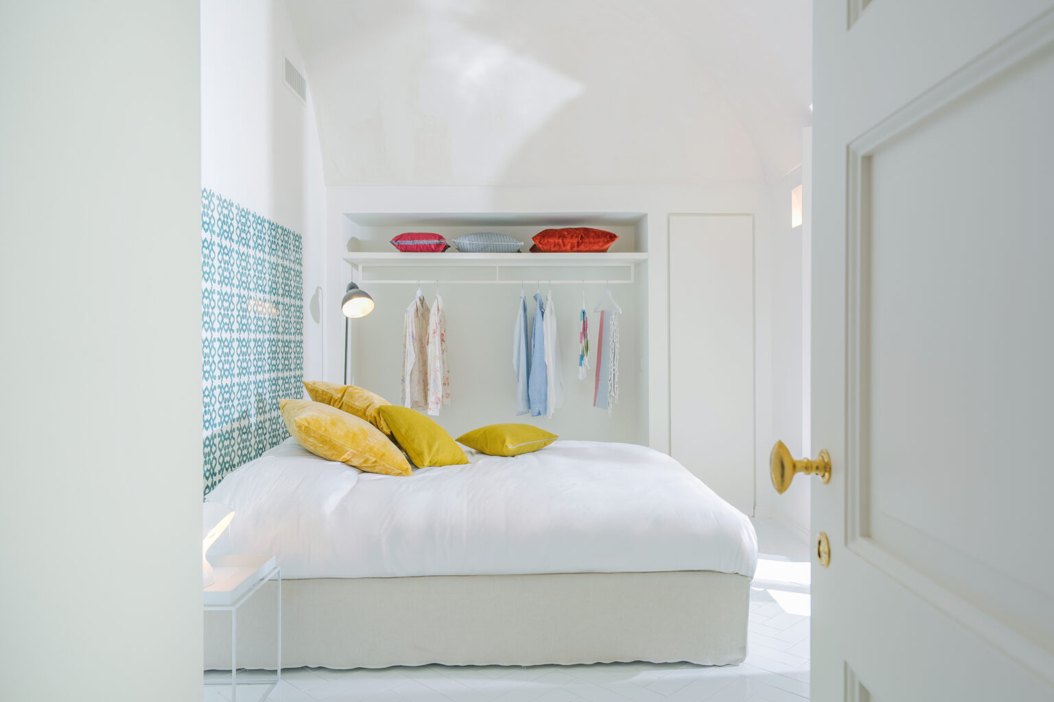 Bright and airy bedroom featuring a large bed with yellow pillows, a stylish hanging wardrobe displaying various garments, and decorative cushions on a shelf. The room has a modern design with white walls and unique patterned tiles, creating a serene atmosphere. | Only on SopranoVillas
