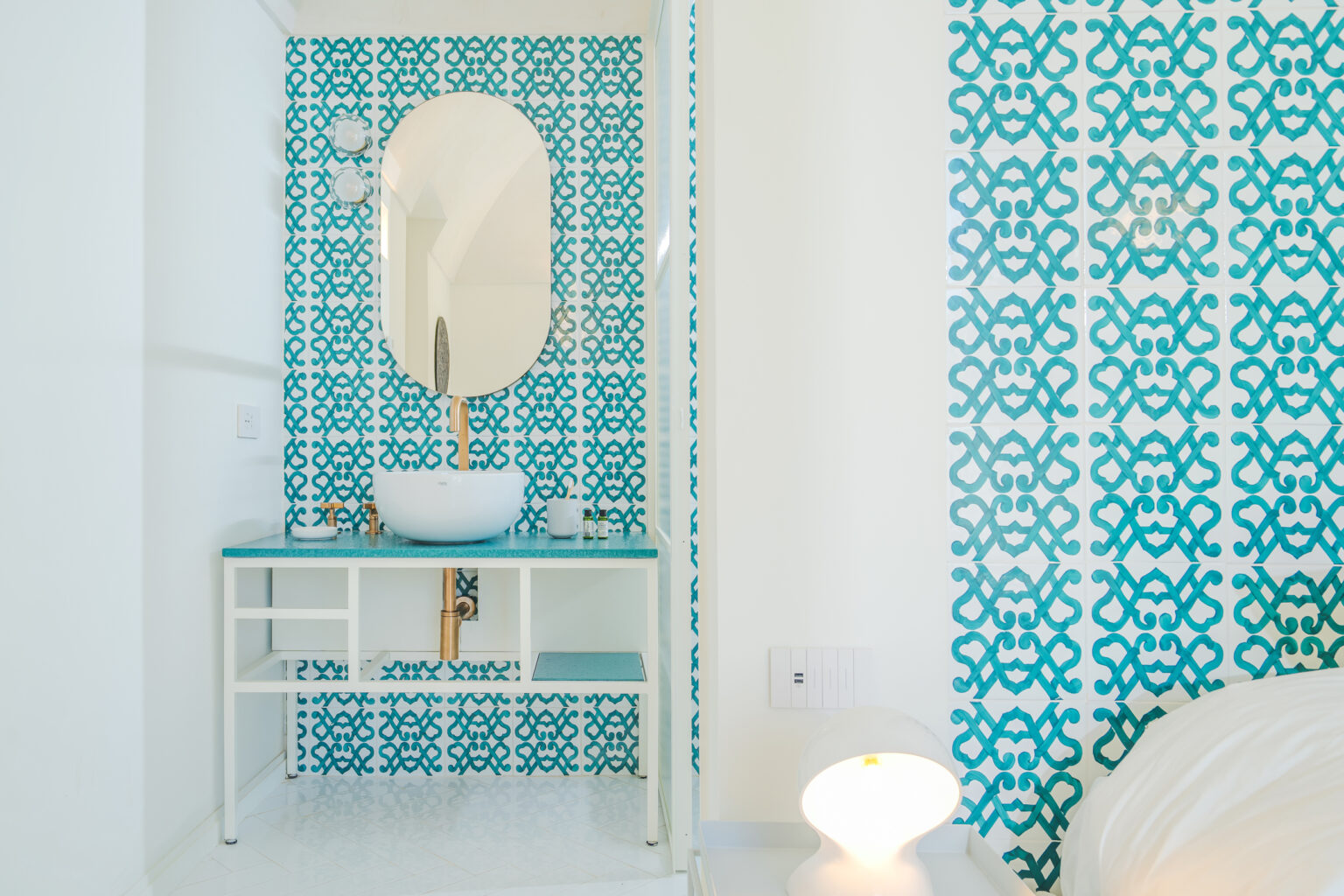 Modern bathroom design featuring a stylish white vessel sink on a turquoise countertop, framed by a decorative wall with intricate blue patterns. The space includes a large oval mirror and elegant lighting, creating a bright and inviting atmosphere. | Only on SopranoVillas