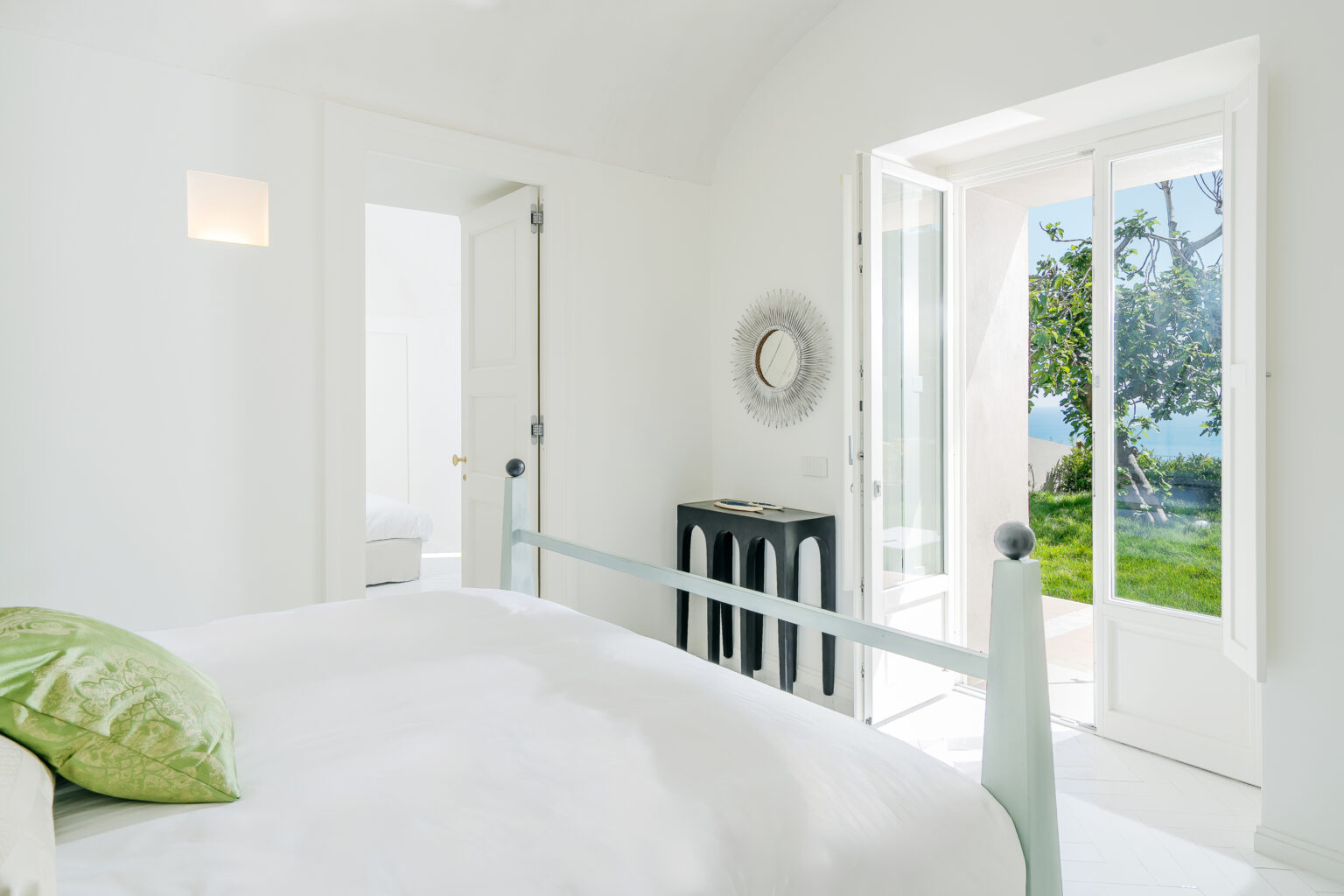Bright and airy bedroom featuring a white color scheme, a light green decorative pillow, and a stylish black console table. The room includes large windows that open to a scenic outdoor view with greenery, enhancing the tranquil atmosphere. | Only on SopranoVillas