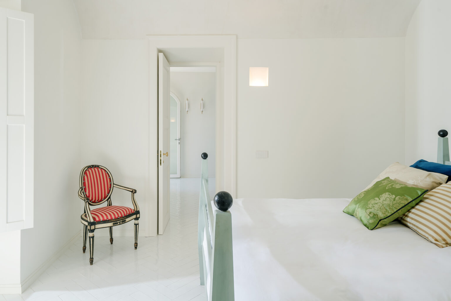 Bright and airy bedroom featuring a stylish bed with decorative pillows and a vintage striped chair. The minimalist design includes white walls and herringbone flooring, creating a serene atmosphere. A doorway leads to another room, enhancing the spacious feel. | Only on SopranoVillas