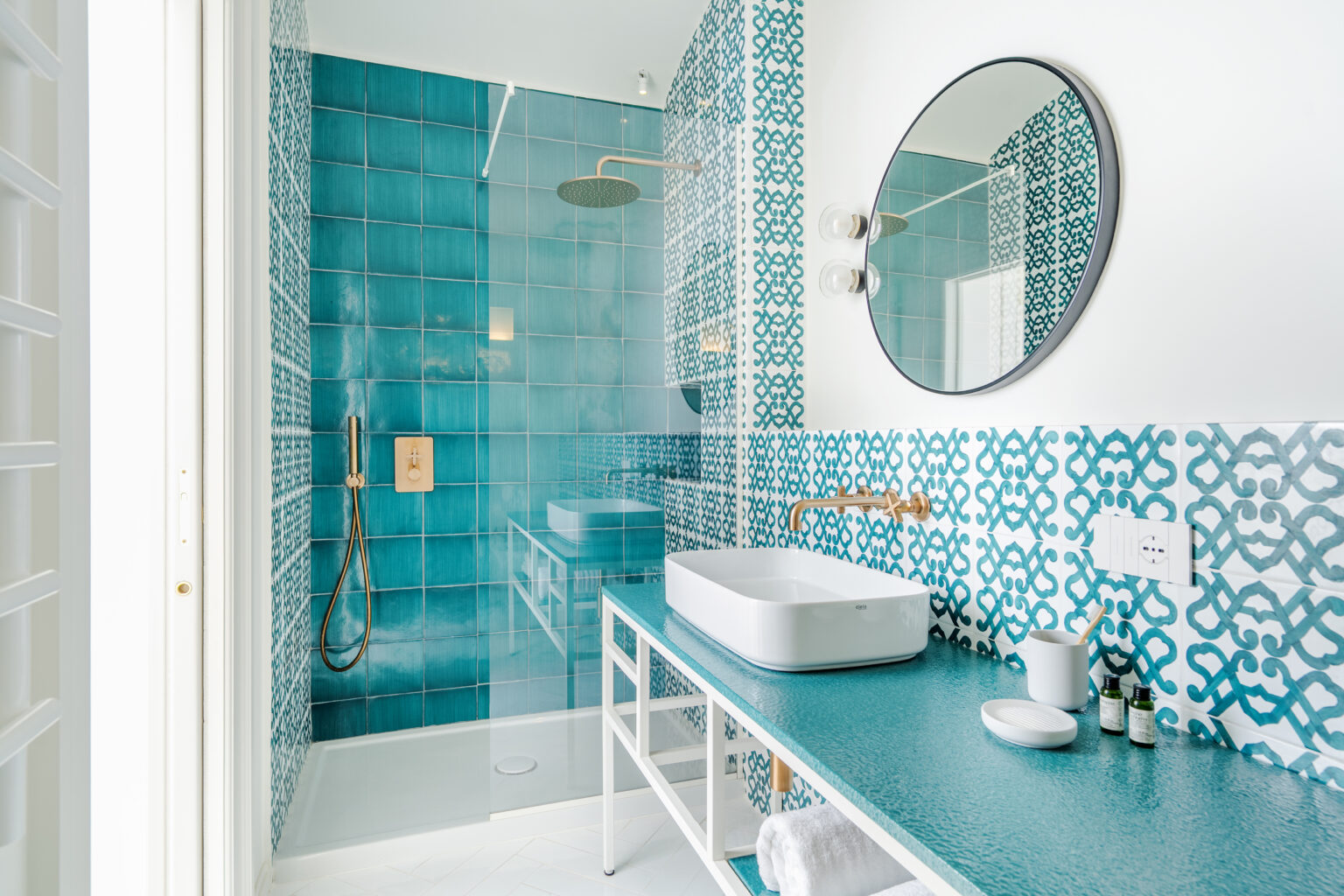 Modern bathroom featuring turquoise tiled walls, a sleek white sink on a matching countertop, and a round mirror. The space includes a glass shower enclosure, stylish fixtures, and decorative elements, creating a fresh and inviting atmosphere. Ideal for home design inspiration. | Only on SopranoVillas