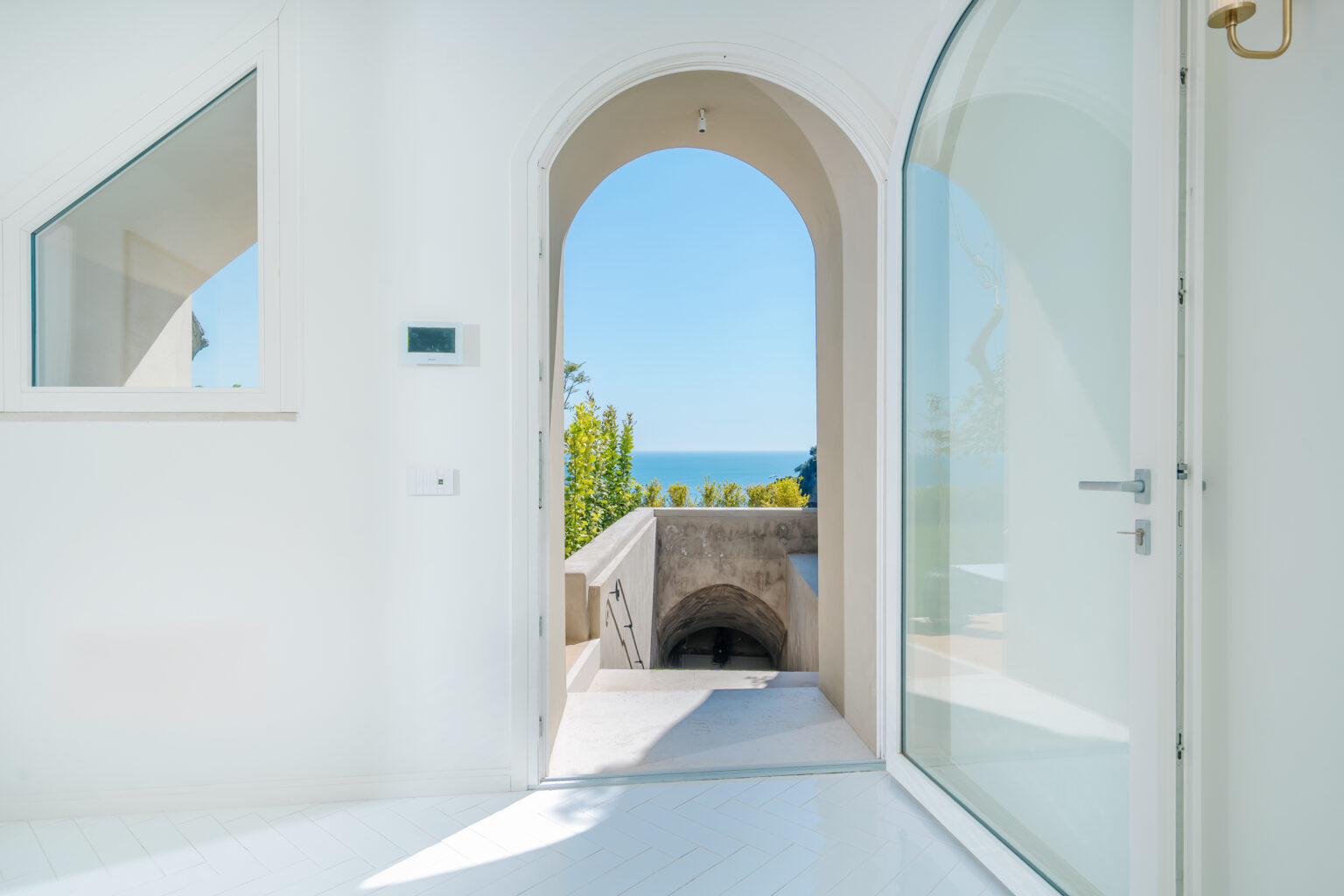 Bright and airy interior view through an arched doorway, leading to a scenic outdoor area with ocean views and lush greenery. The space features modern design elements, including large glass doors and white walls, enhancing the natural light. | Only on SopranoVillas