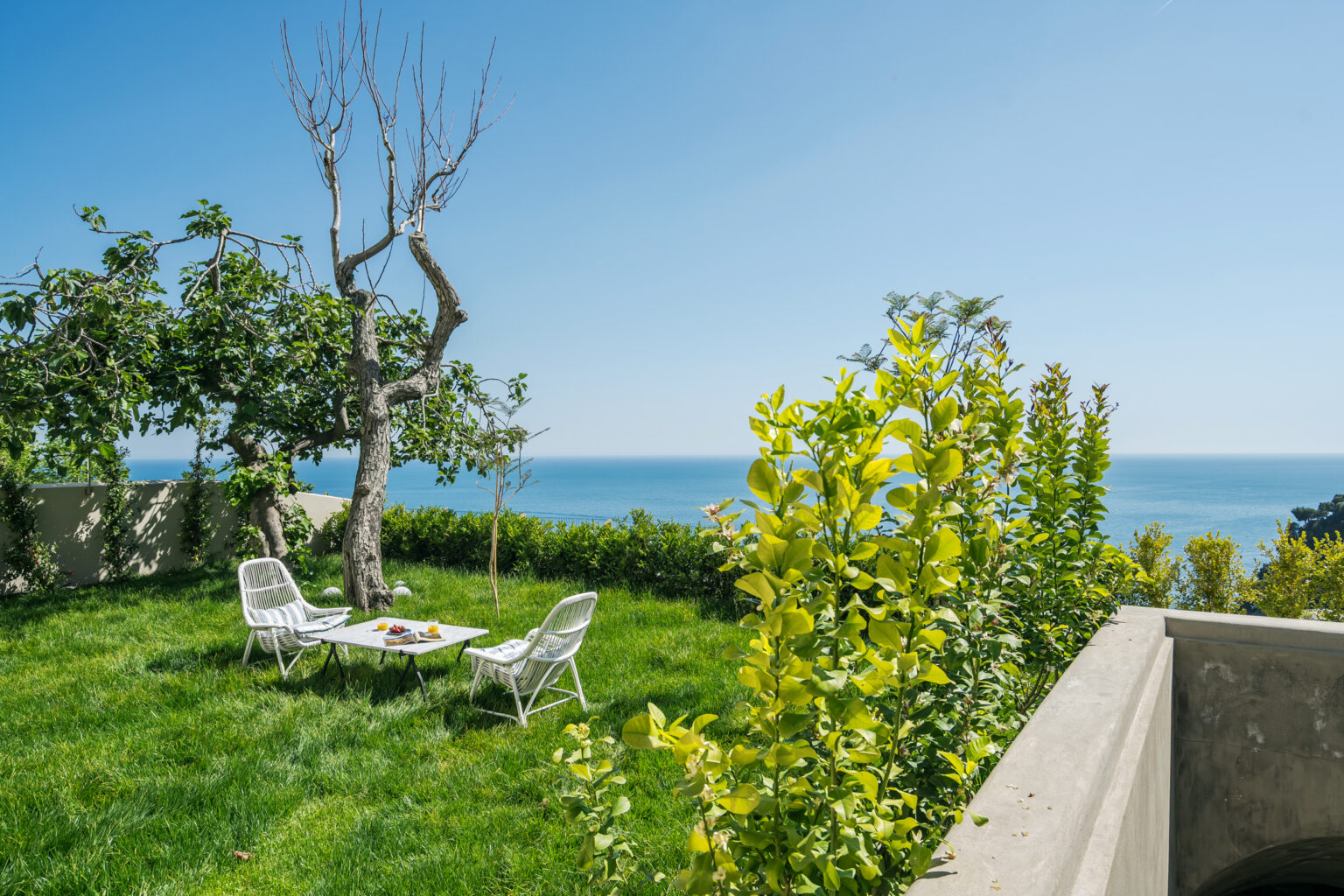 Cozy outdoor seating area with white chairs and a table, surrounded by lush green grass and vibrant foliage, overlooking a serene ocean view under a clear blue sky. | Only on SopranoVillas