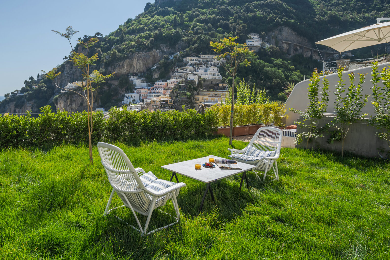 Cozy outdoor seating area with two white chairs and a marble table, surrounded by lush green grass and a scenic view of hillside homes and trees. Ideal for relaxation and enjoying the landscape. | Only on SopranoVillas