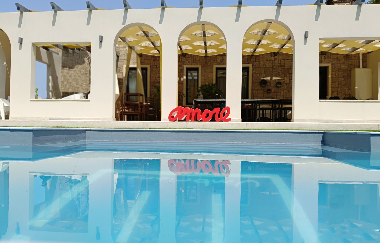 Vibrant red "amore" sign in front of a modern villa with arched openings, reflecting in a serene blue swimming pool under a clear sky. | Only on SopranoVillas