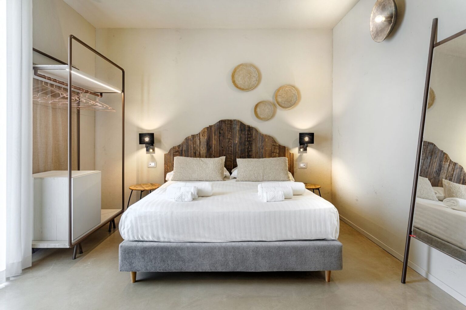 Modern hotel room featuring a cozy double bed with a wooden headboard, two decorative pillows, and neatly arranged towels. The space includes contemporary decor, a standing mirror, minimalistic clothing rack, soft lighting, and a warm color palette, creating a comfortable and inviting atmosphere. | Only on SopranoVillas