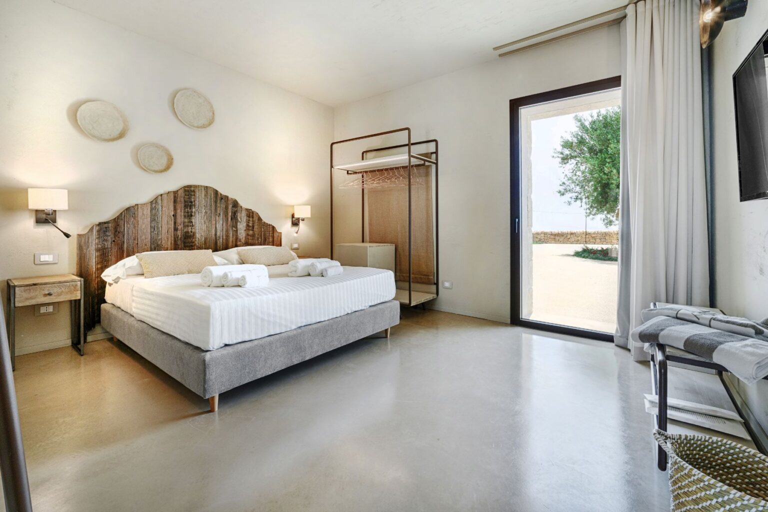 Contemporary hotel bedroom featuring a rustic wooden headboard, plush bedding, and modern amenities. The room includes a large window with outdoor views, a stylish clothing rack, and soft lighting, creating a cozy and inviting atmosphere. Perfect for relaxing retreats or business travel. | Only on SopranoVillas