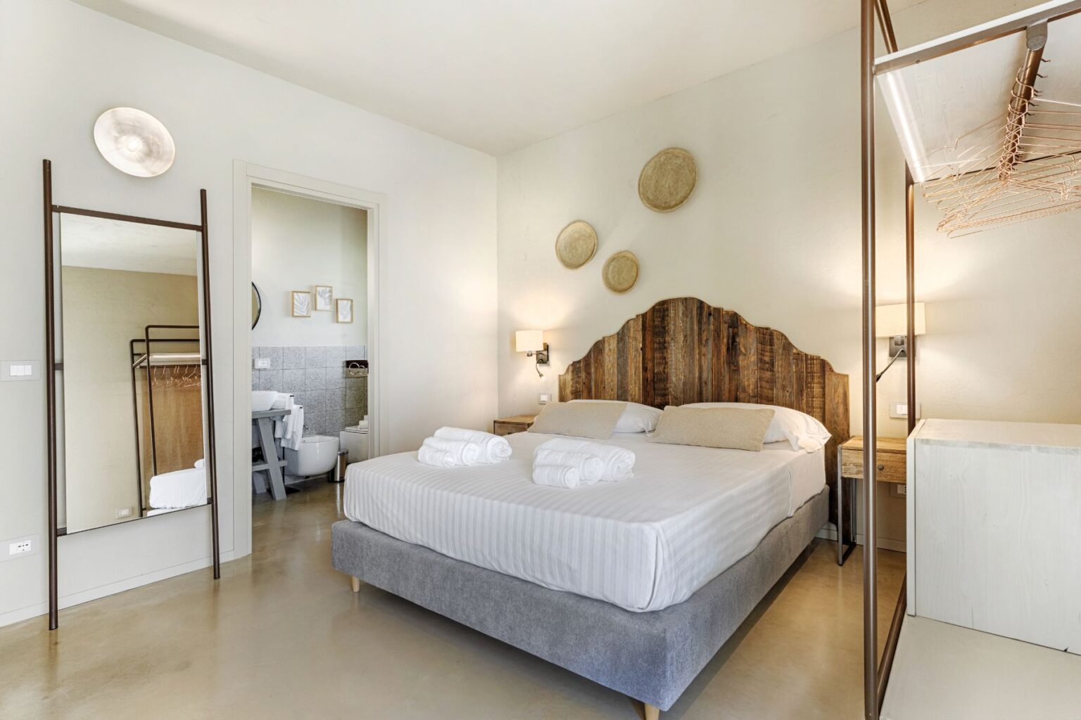 Modern hotel room featuring a stylish wooden headboard, plush bed with rolled towels, a full-length mirror, and a compact bathroom visible through an adjoining door. | Only on SopranoVillas