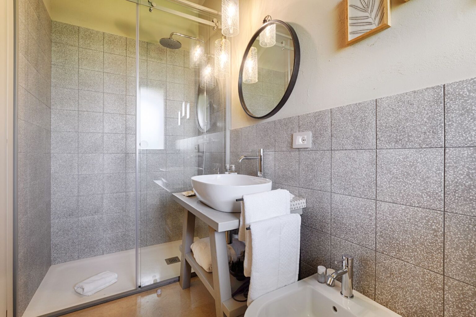 Modern bathroom featuring a walk-in shower with glass doors, a stylish round mirror, a sleek white sink on a wooden vanity, and neatly arranged towels. The gray tiled walls and hanging pendant lights create a contemporary and inviting atmosphere. | Only on SopranoVillas