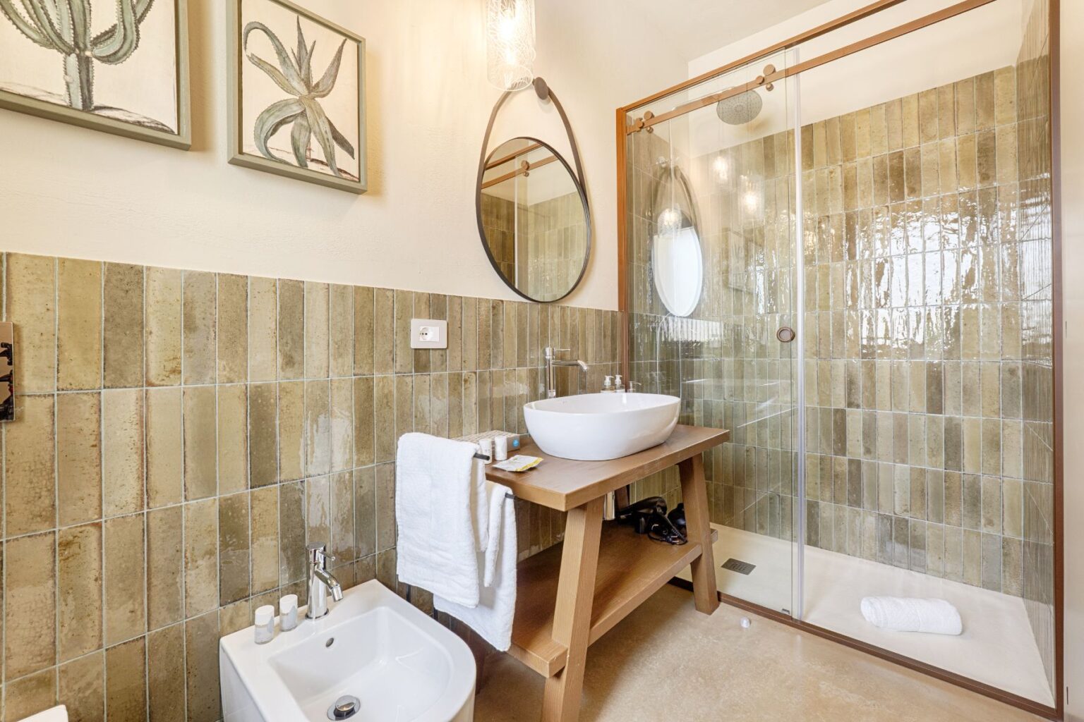 Modern bathroom interior featuring a stylish shower with glass enclosure, a round mirror, and a wooden vanity with a white vessel sink. Natural stone tiles create an earthy ambiance, complemented by decorative wall art of cacti. Soft towels and toiletries are neatly arranged for convenience. | Only on SopranoVillas