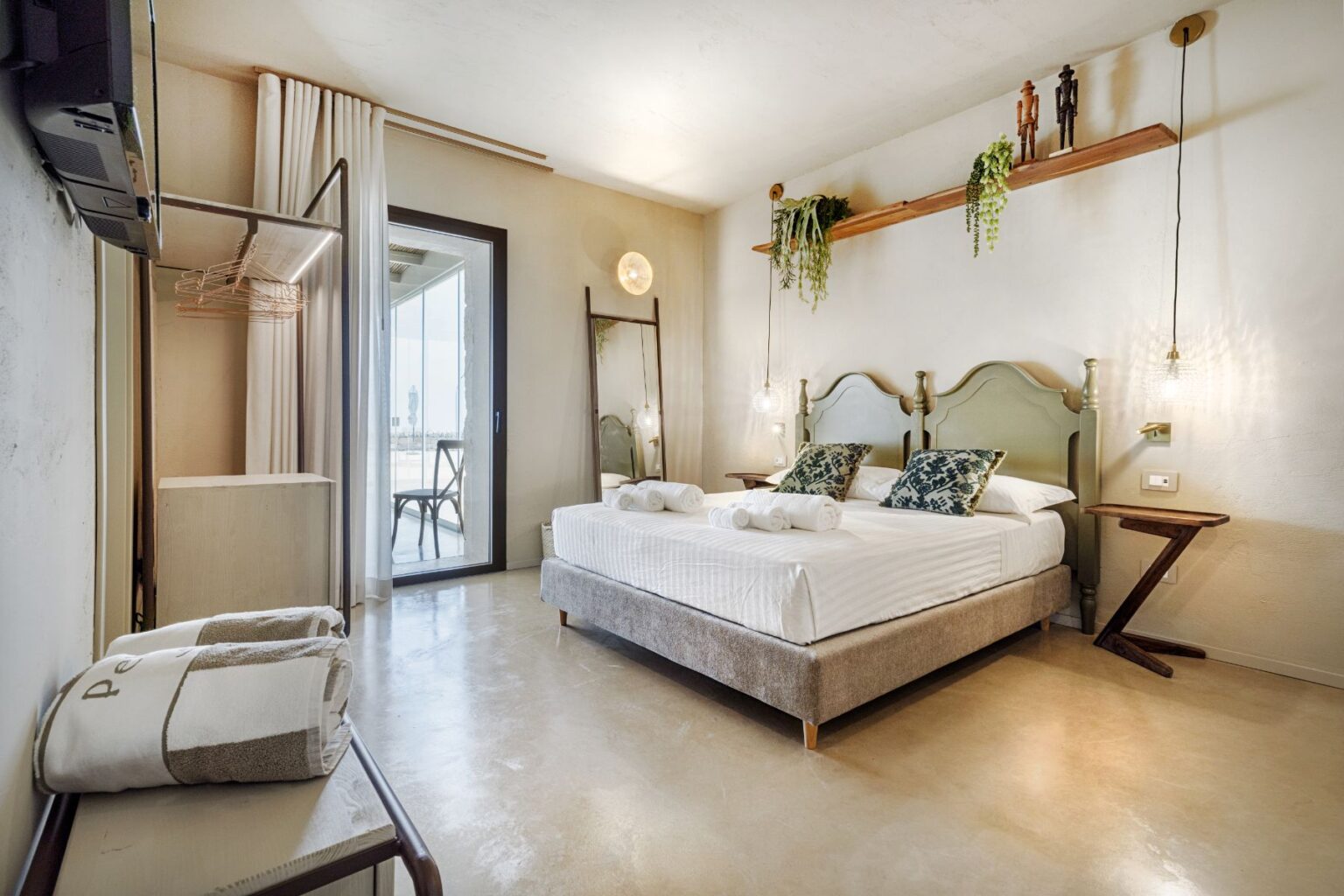 Modern hotel room featuring a comfortable double bed with decorative pillows, rolled towels, a seating area, and large windows that open to a balcony. The room is styled with soft lighting, wooden decor accents, and greenery, creating a cozy and inviting atmosphere. | Only on SopranoVillas