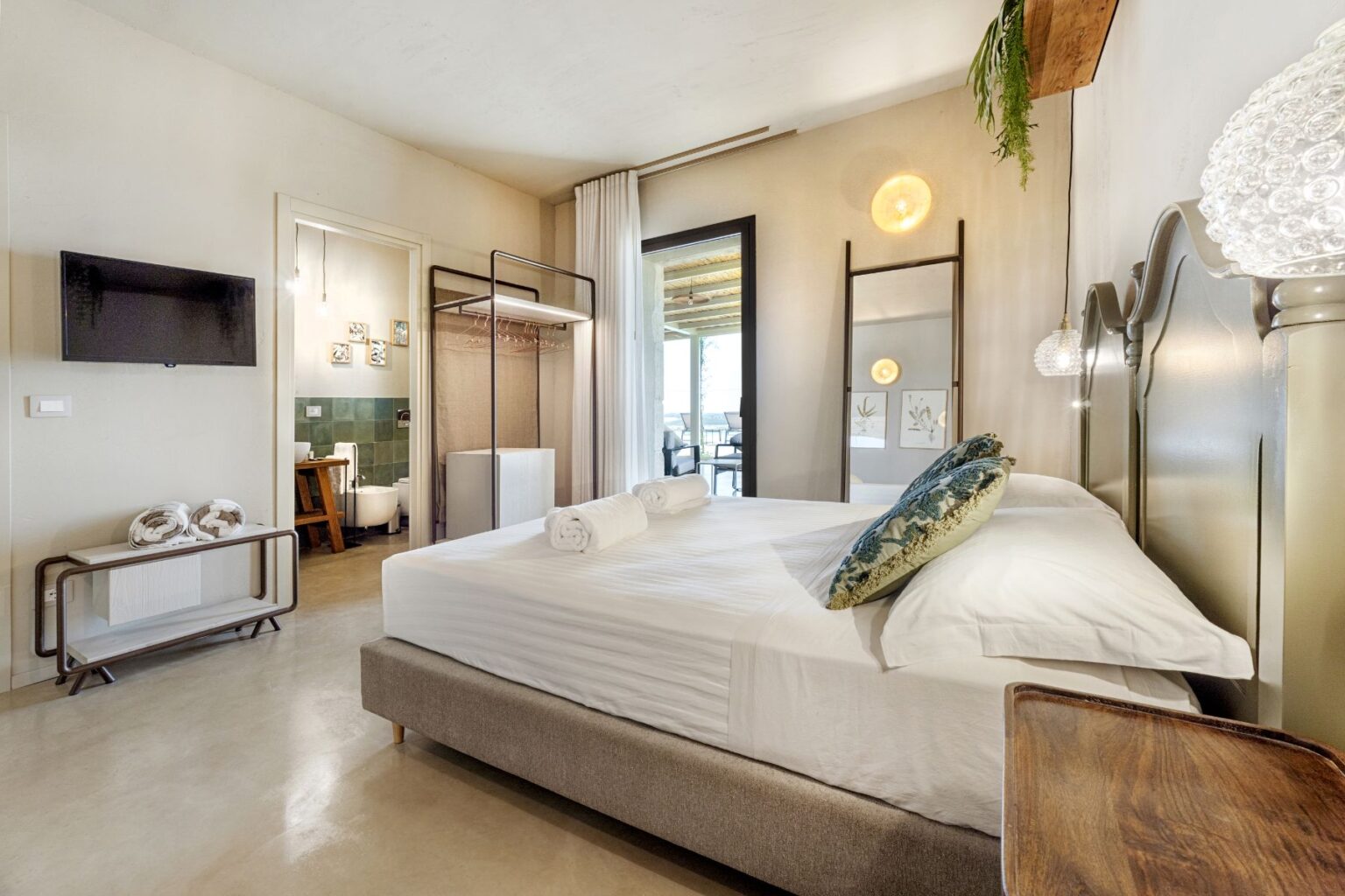 Stylish hotel room featuring a plush bed with decorative pillows, modern decor, and a wall-mounted TV. The space includes an open closet area, an en-suite bathroom, and large windows that offer natural light. Ideal for relaxation and comfort. | Only on SopranoVillas