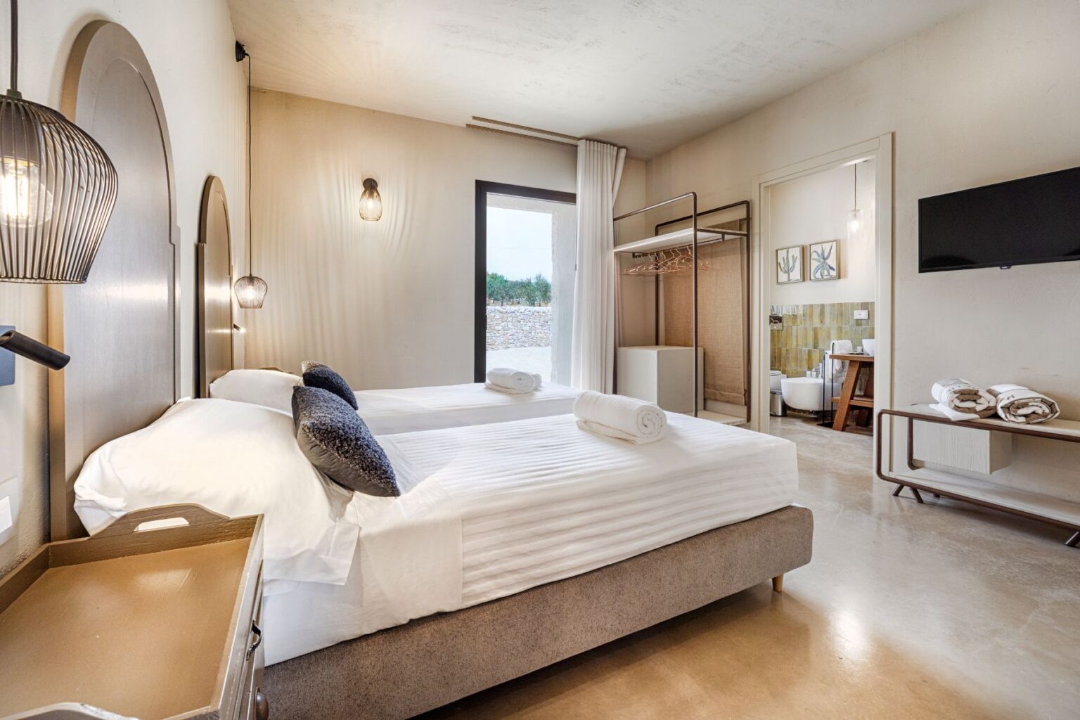 Modern hotel room with two twin beds, decorative pillows, and soft lighting. Features a minimalist design with a wardrobe, TV, and an en-suite bathroom visible through an open door. Natural light streams in from a window, adding warmth to the serene ambiance. Ideal for travelers seeking comfort and style. | Only on SopranoVillas