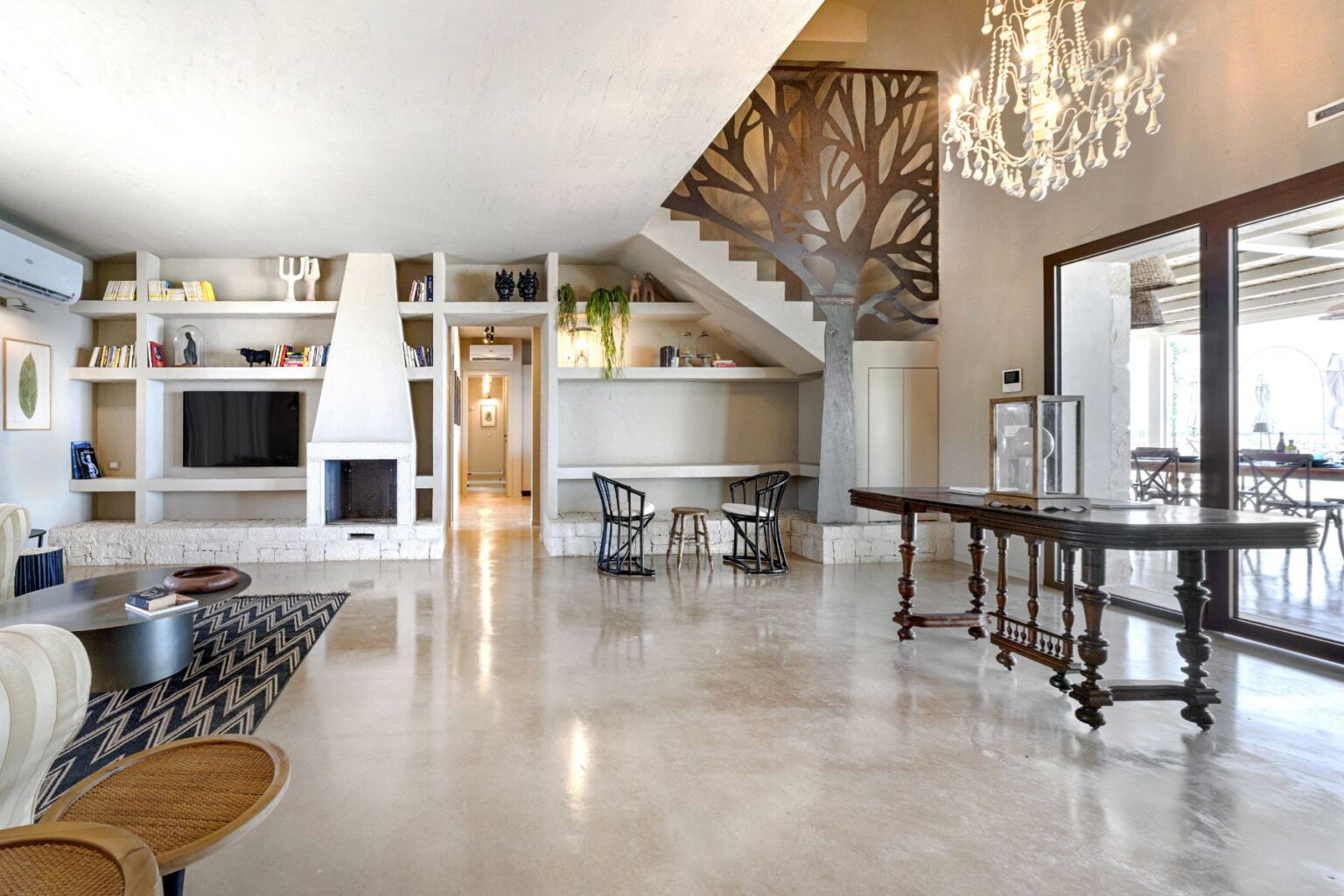 Spacious modern living room featuring high ceilings, a unique wall design, built-in shelves with books, a central fireplace, stylish furniture, and a large window providing natural light. | Only on SopranoVillas