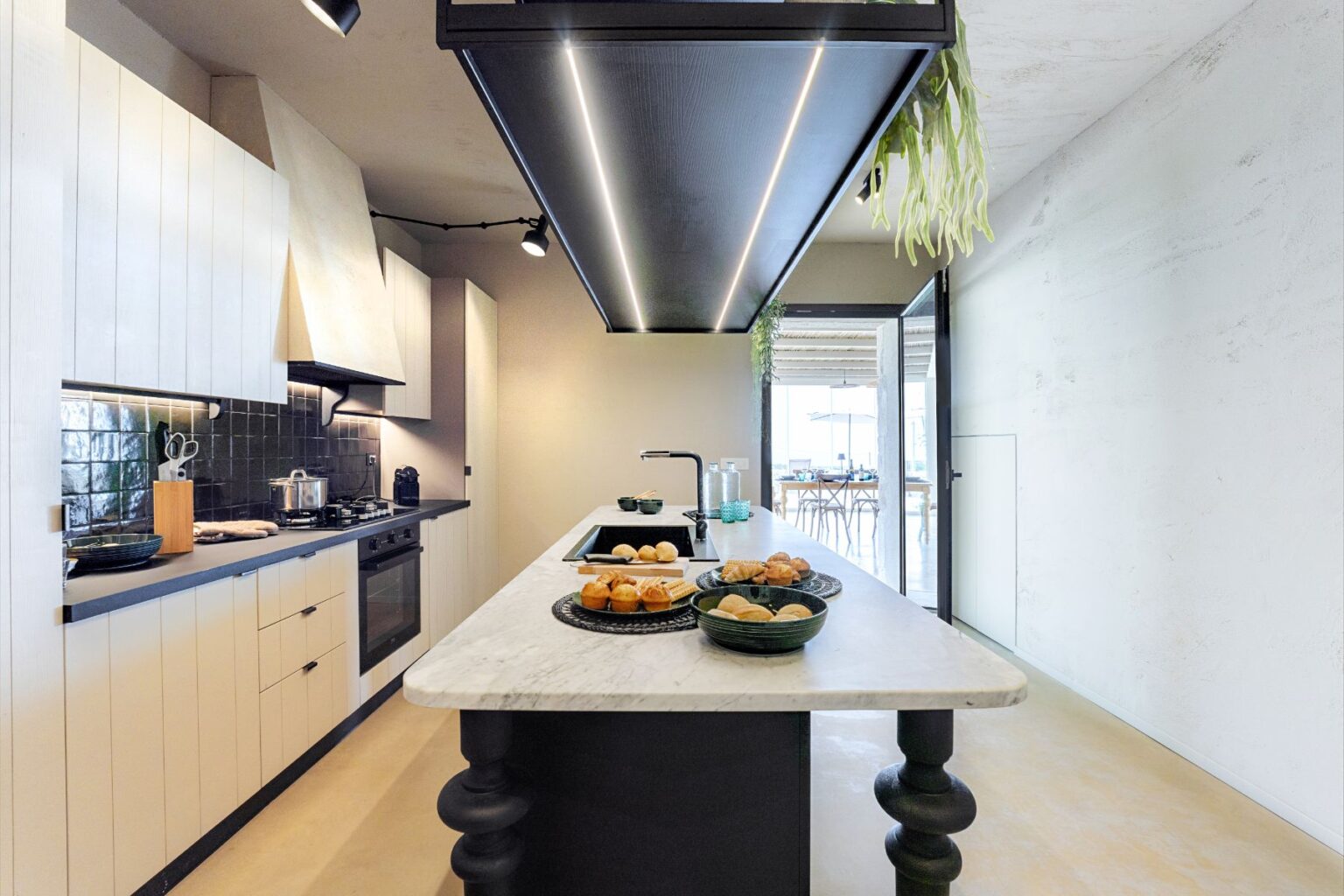 Modern kitchen with marble island, black and white cabinetry, and stylish appliances, featuring an array of pastries on the counter, and large windows allowing natural light to fill the space. | Only on SopranoVillas