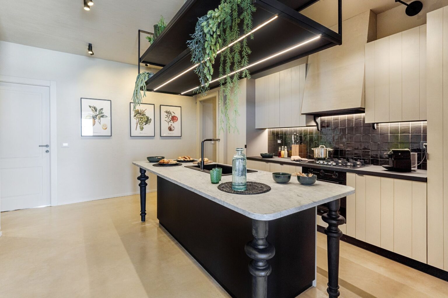 Modern kitchen featuring a central island with a marble countertop, stylish black cabinetry, and decorative wall art of fruits and herbs. The space is illuminated by overhead lighting and lush greenery hangs from a shelf, creating a fresh and inviting atmosphere. | Only on SopranoVillas