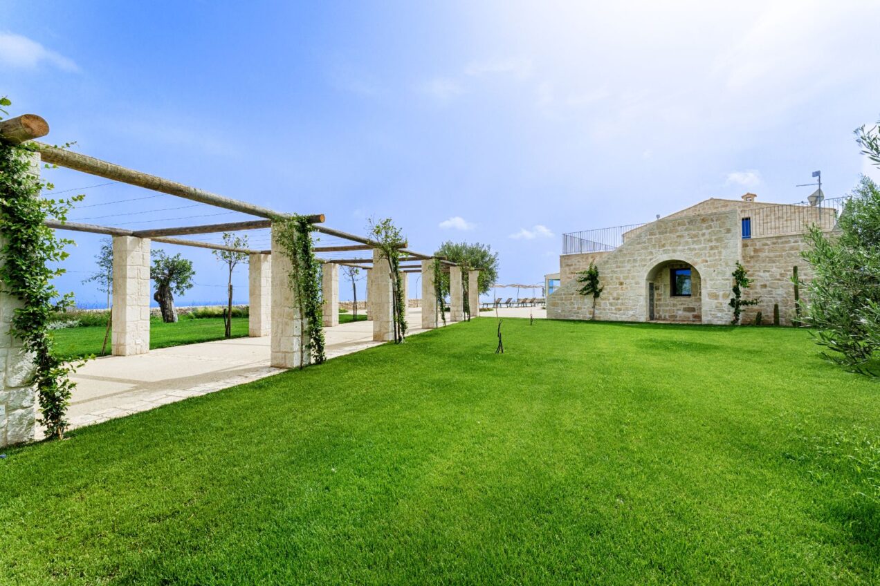 Beautiful landscaped garden with stone pathways and trellises, featuring lush green grass and blue skies in the background, showcasing a charming architectural structure. Ideal for outdoor gatherings and relaxation. | Only on SopranoVillas