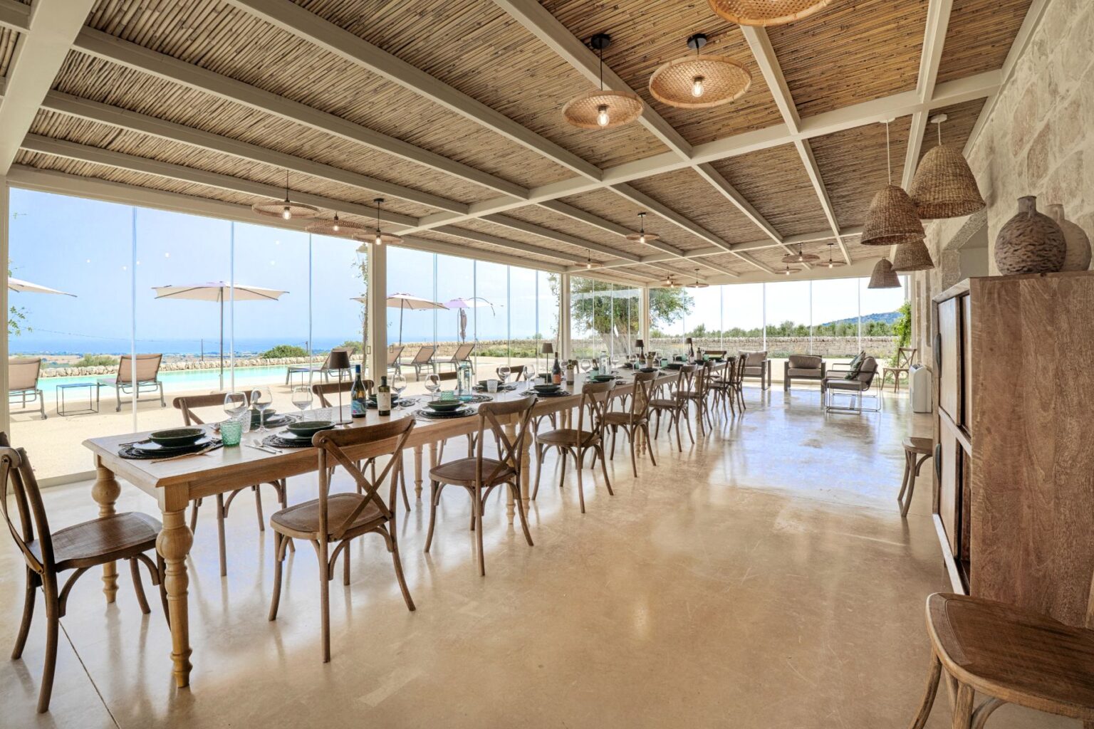 Spacious modern restaurant interior showcasing a long wooden dining table set for a meal, with elegant tableware and bottles of wine. Large glass windows offer a panoramic view of the outdoor pool and landscape, enhancing the bright, airy atmosphere. Stylish hanging lights and woven decor elements add warmth to the design. | Only on SopranoVillas