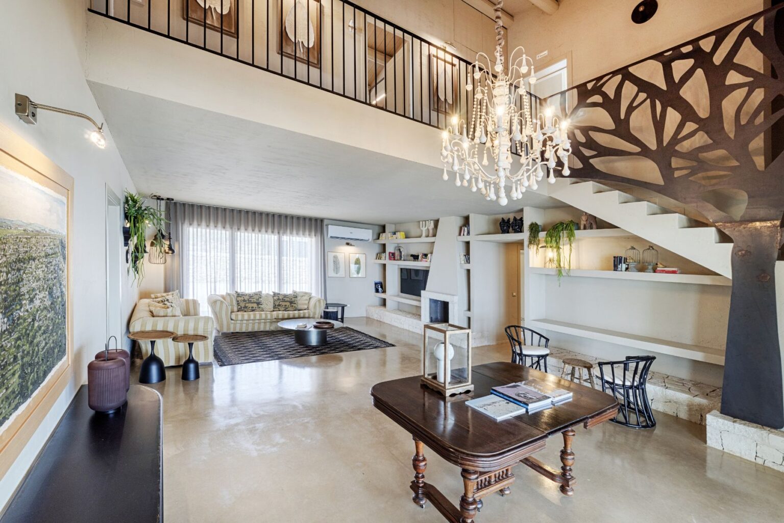 Spacious modern living room featuring a beige striped sofa, a hardwood table, and a stylish chandelier. The interior showcases large windows with sheer curtains, polished concrete floors, and decorative wall art, creating a bright and inviting atmosphere. A staircase with a unique railing design leads to an upper level, enhancing the contemporary aesthetic. | Only on SopranoVillas