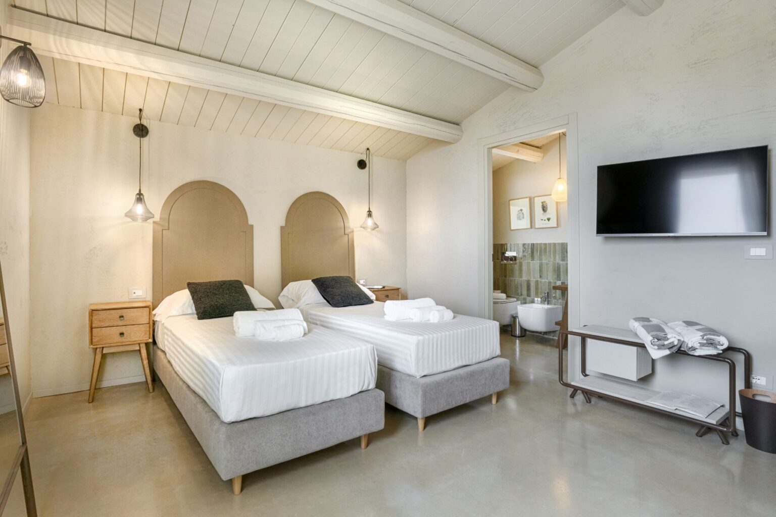 Modern hotel room featuring two twin beds with plush bedding, soft pillows, and neatly rolled towels. The decor includes a minimalist design with wooden accents, stylish pendant lighting, and a flat-screen TV. An open doorway reveals a chic bathroom area, enhancing the room's spacious feel. Perfect for travelers seeking comfort and elegance. | Only on SopranoVillas