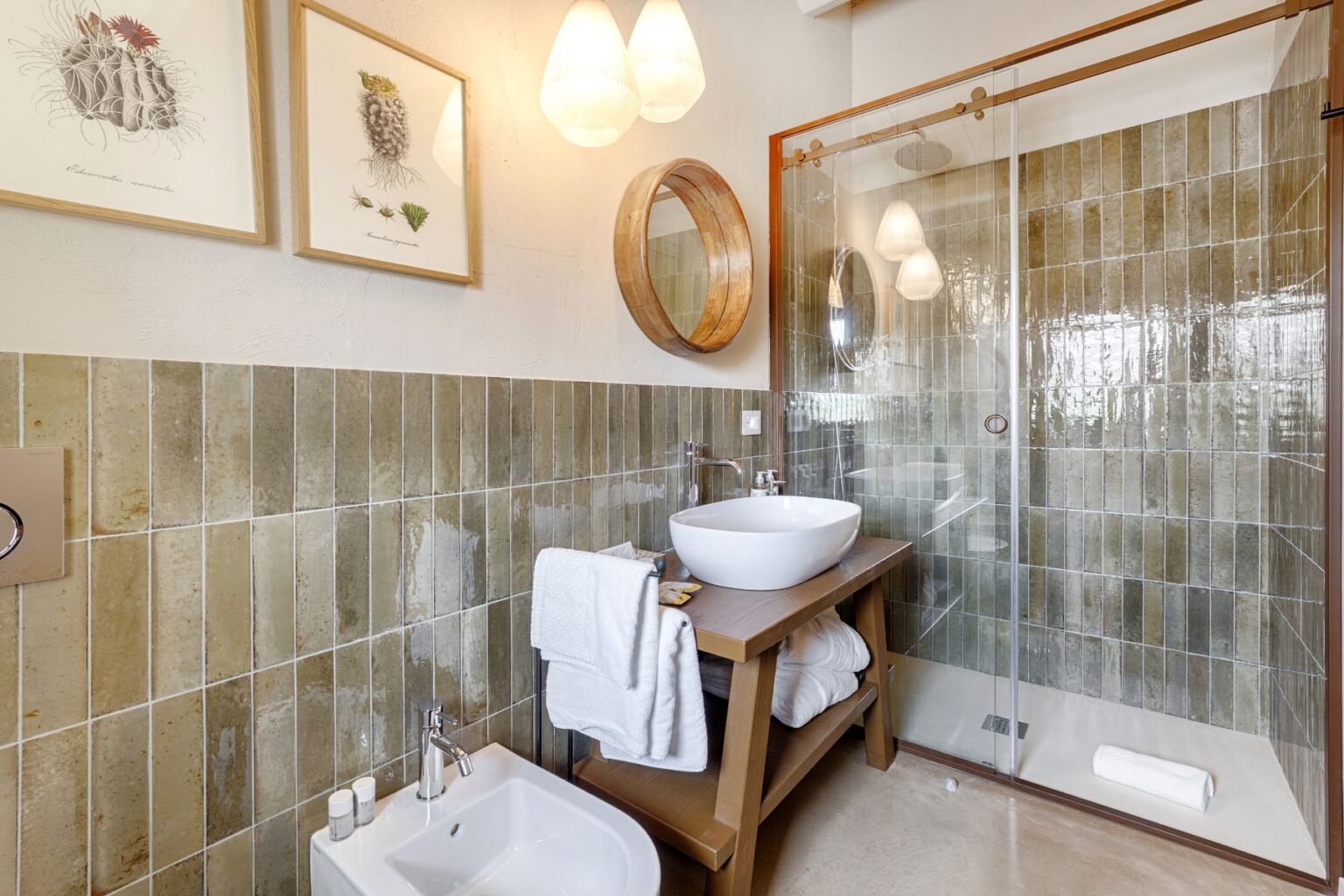 Modern bathroom interior featuring a glass shower, ceramic sink on a wooden vanity, and stylish wall tiles. Decor includes framed botanical prints and soft lighting, creating a chic and inviting atmosphere. | Only on SopranoVillas