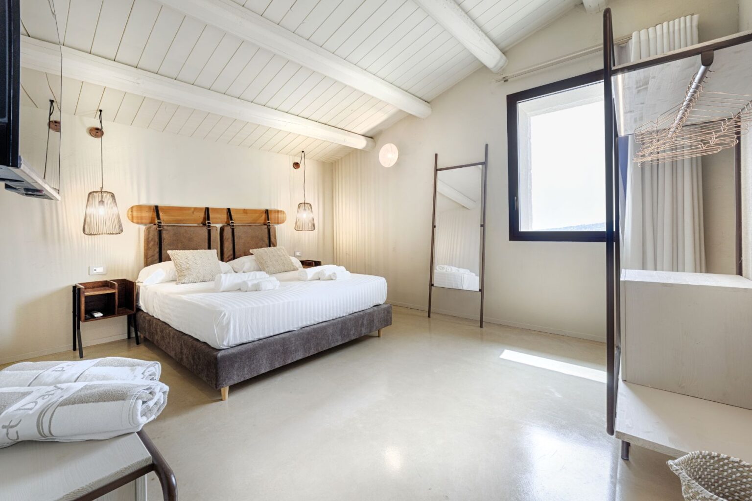 Modern and stylish hotel room featuring a double bed with decorative pillows, minimalist decor, natural light from a large window, and contemporary lighting fixtures. Amenities include a mirror and a wooden shelf for storage, ideal for a comfortable stay. | Only on SopranoVillas