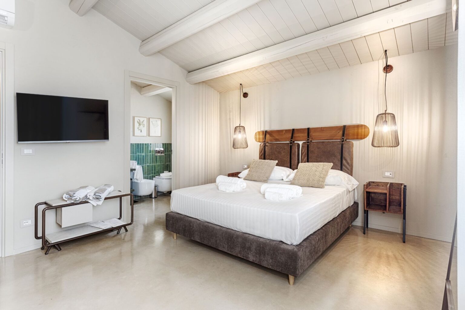 Spacious and modern hotel room featuring a large double bed with stylish linen, a flat-screen TV, and decorative lighting. The room is designed with neutral tones and includes a small bedside table. A glimpse of the en-suite bathroom with green tiled walls is visible in the background. Ideal for a comfortable stay. | Only on SopranoVillas